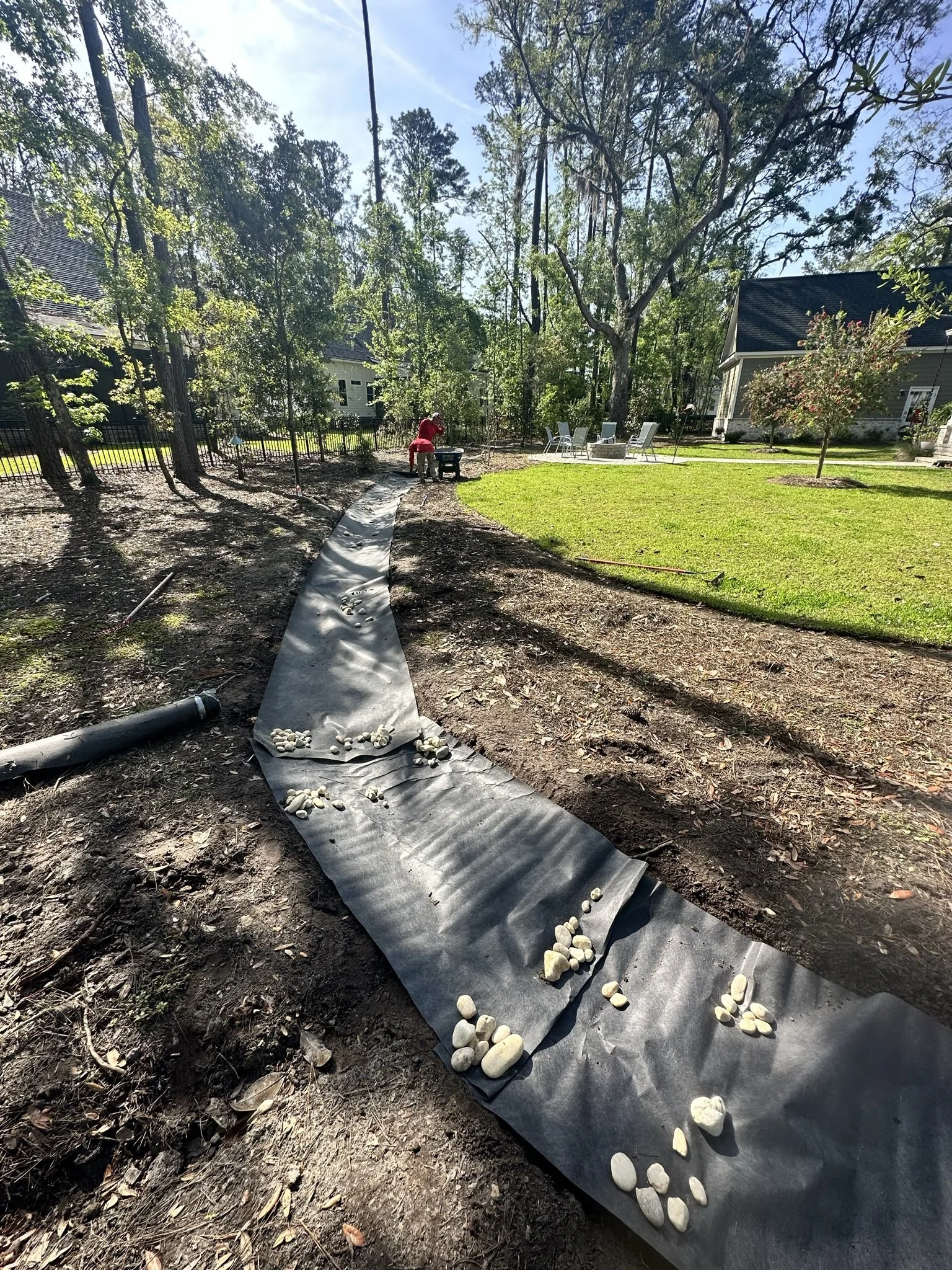 A backyard with a curved path under construction, with stones laid out along the pathway, surrounded by trees, grass, and some outdoor furniture, with a person working in the background.