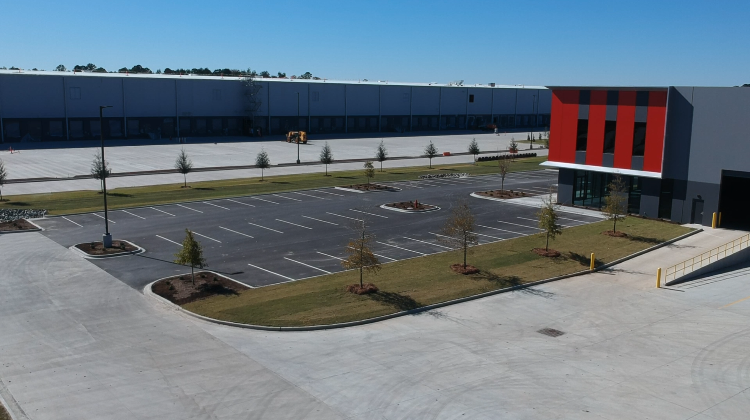 Empty parking lot in front of a large commercial building with red and gray exterior, small trees, and a clear blue sky.