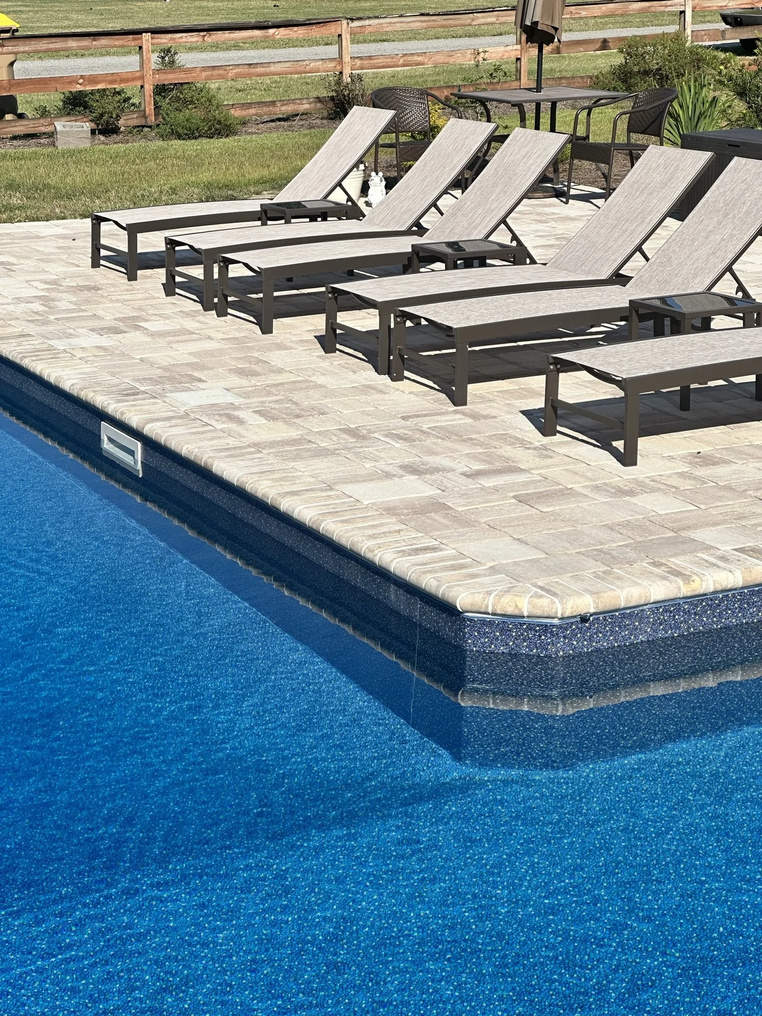 Poolside scene with six lounge chairs on a stone patio, a small table and chairs in the background, enclosed by a wooden fence and some greenery.
