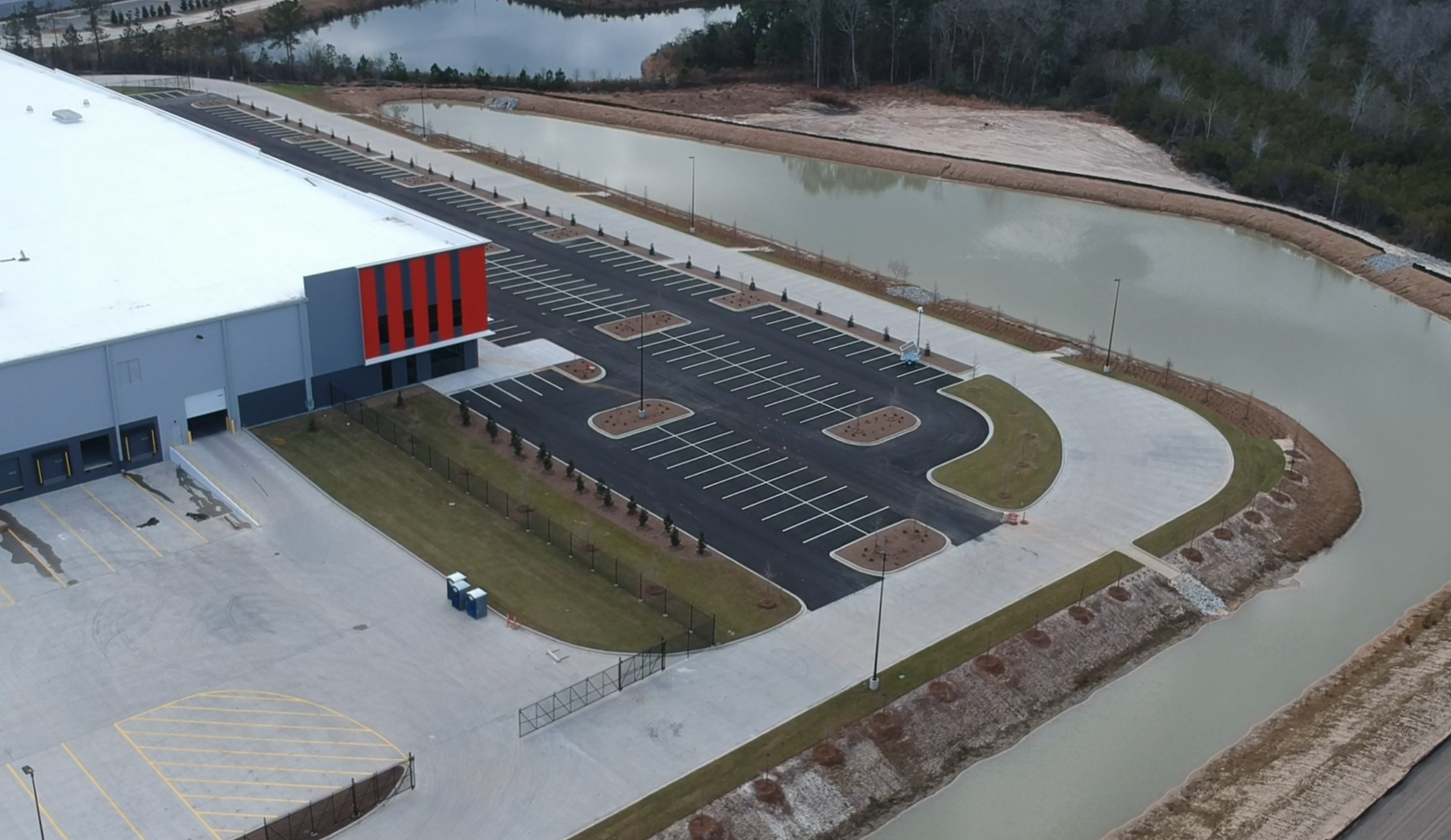 Empty parking lot next to a large warehouse with red and gray exterior, surrounded by landscaped areas and adjacent to a waterway, with an overcast sky in the background.