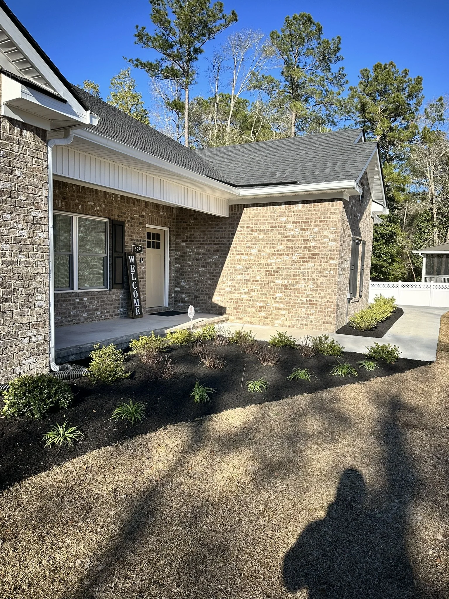 Front porch of a brick house with a welcome sign, landscaped yard with small bushes and plants, concrete walkway, and a blue sky with green trees in the background.