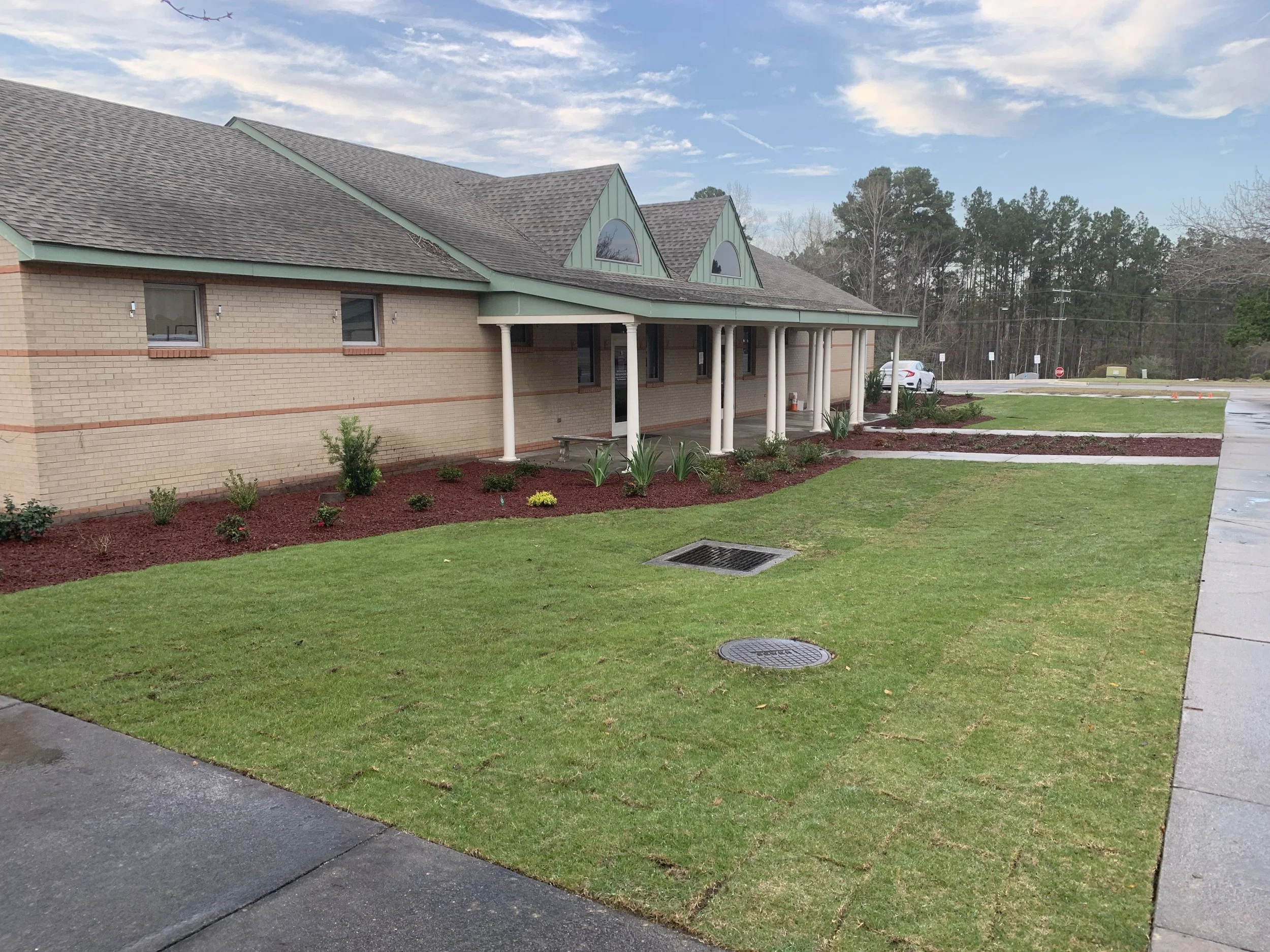 A single-story brick building with a covered porch supported by white columns, surrounded by a well-maintained lawn and flower beds with small plants and bushes.