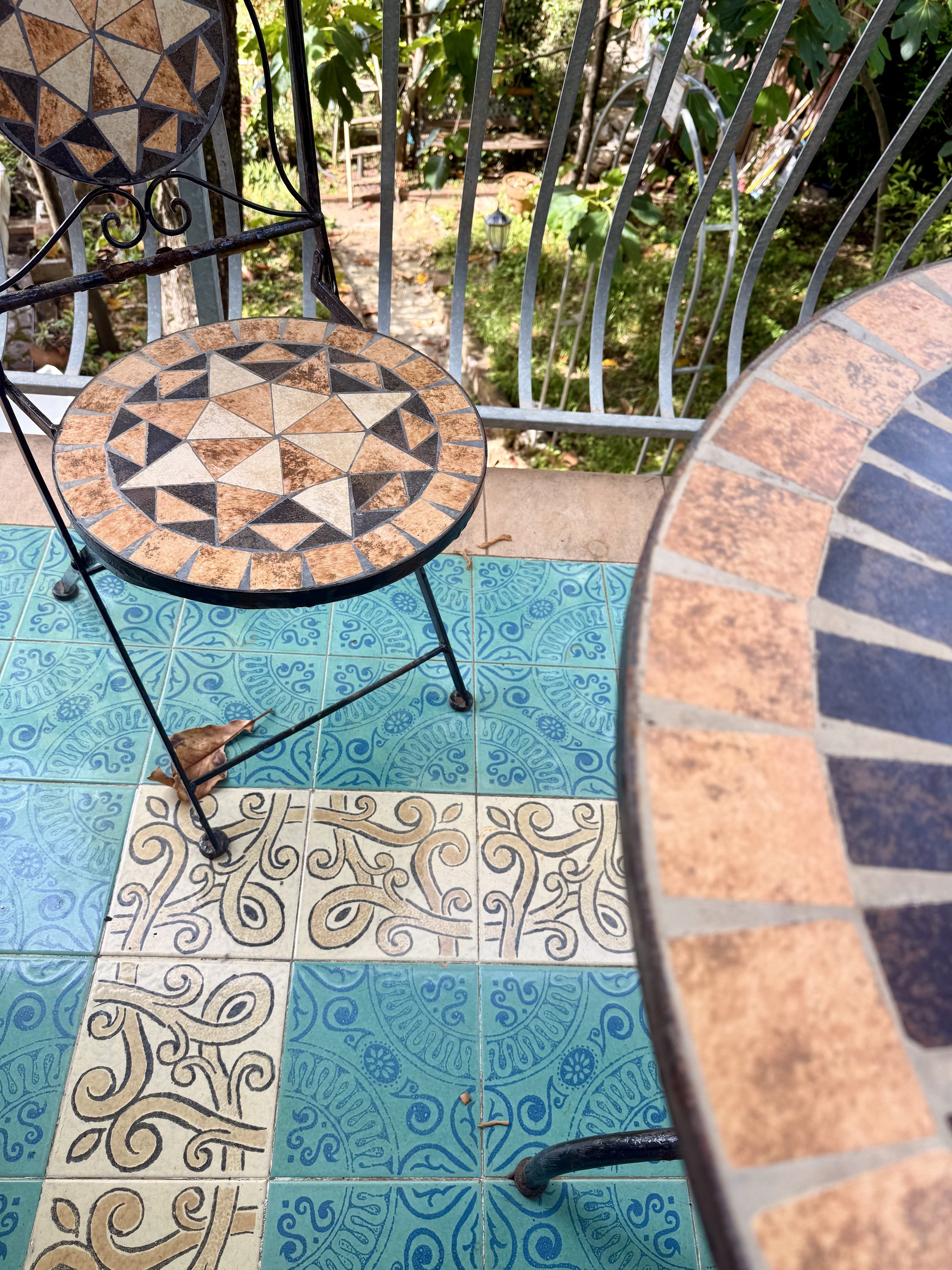 A small round table with a mosaic tile top, sitting on a tiled patio. The mosaic features geometric patterns in brown, black, and beige. Next to the table is a black metal chair with a matching mosaic seat and a decorative backrest. The patio tiles a