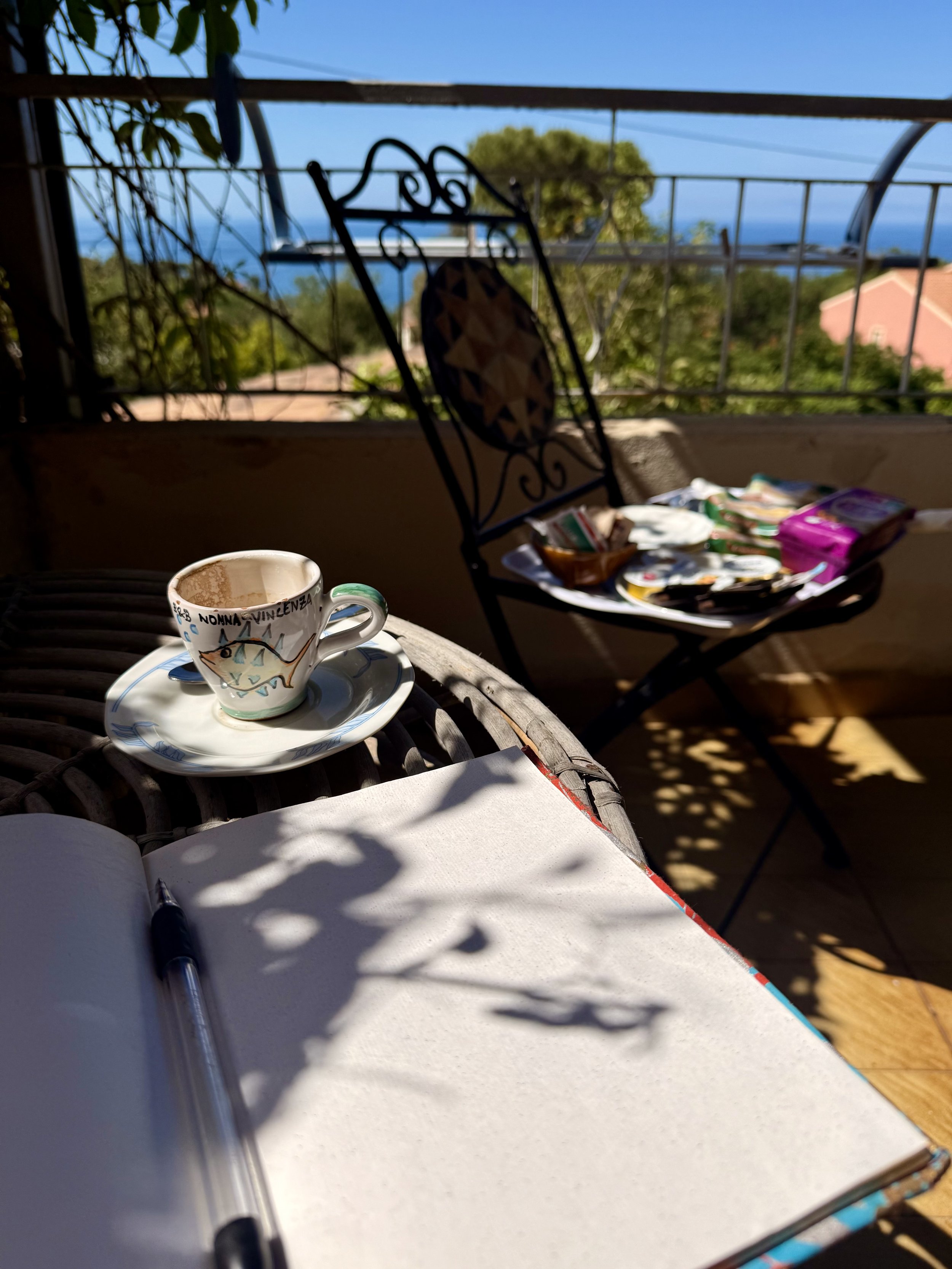 A balcony with a coffee cup on a small table, an open notebook with a pen, and a chair with a tray of snacks in front of a view of trees and the ocean under a clear blue sky.