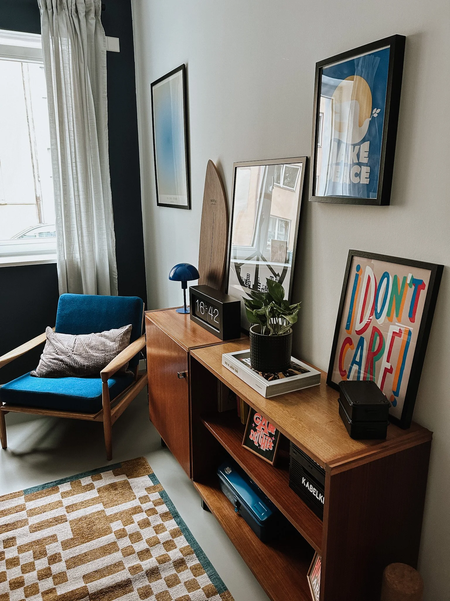 Ein Wohnzimmer mit einer blauen Couch, einem Holztisch mit Uhr, Pflanzen, Bildern an der Wand, einem Fenster mit weißen Vorhängen und einem Teppich mit braunen und weißen Mustern.