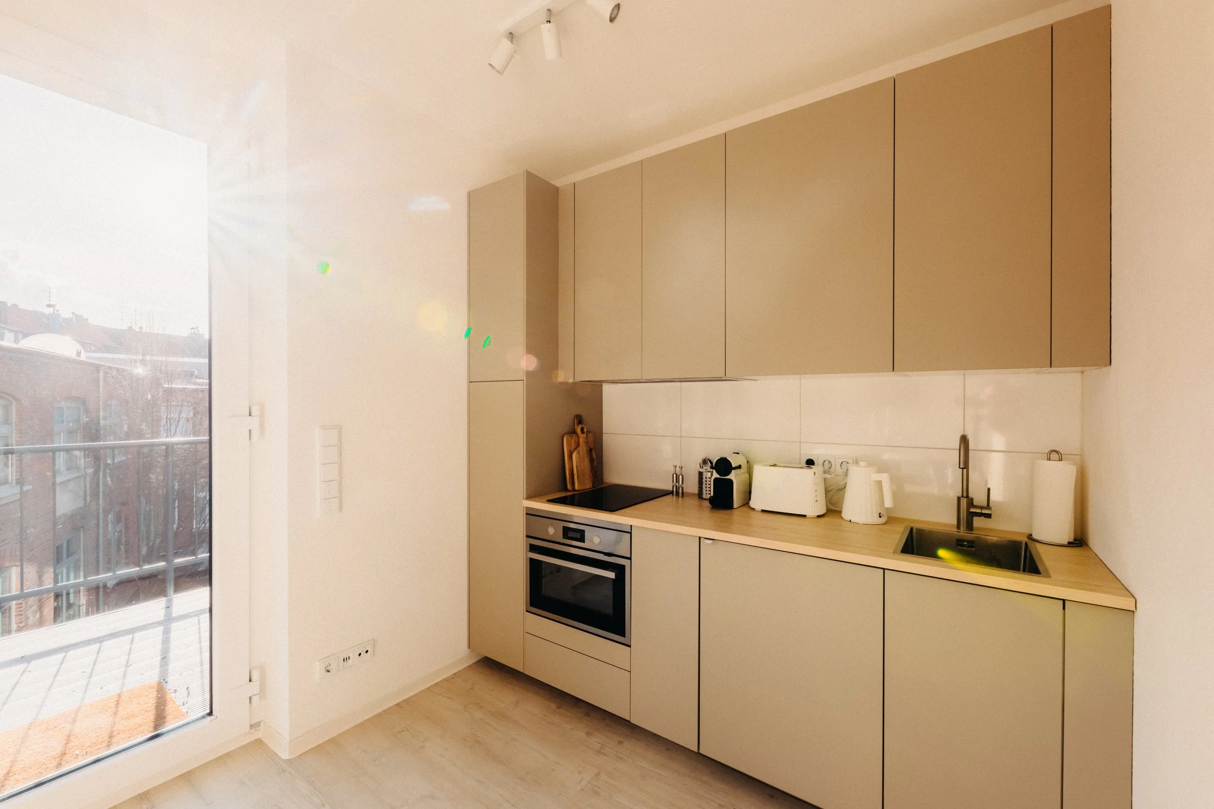 Moderner Einbauküche mit weißen Schränken und Holzarbeitsplatte, darunter Backofen, Herd und Spüle, neben großen Fenstern mit Balkon, die Tageslicht in den Raum lassen.