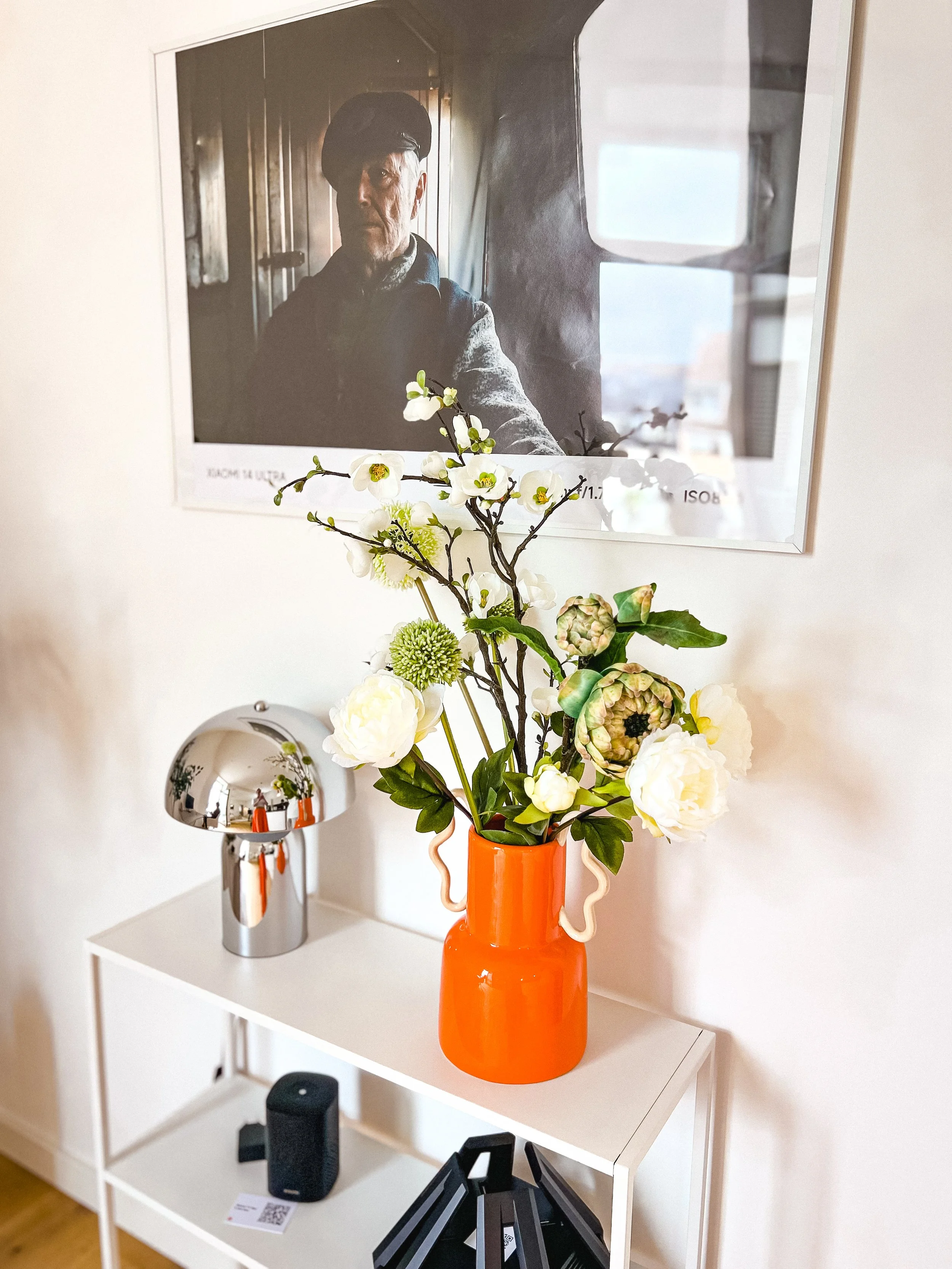Ein weißer Regal mit Blumenstrauß in einer orangefarbigen Vase, einem schwarzen Objekt und einem schrägegelegten Kamera- oder Präsentationsgerät. An der Wand hängt ein gerahmtes Schwarz-Weiß-Foto eines Mannes in dunkler Kleidung und Baskenmütze.