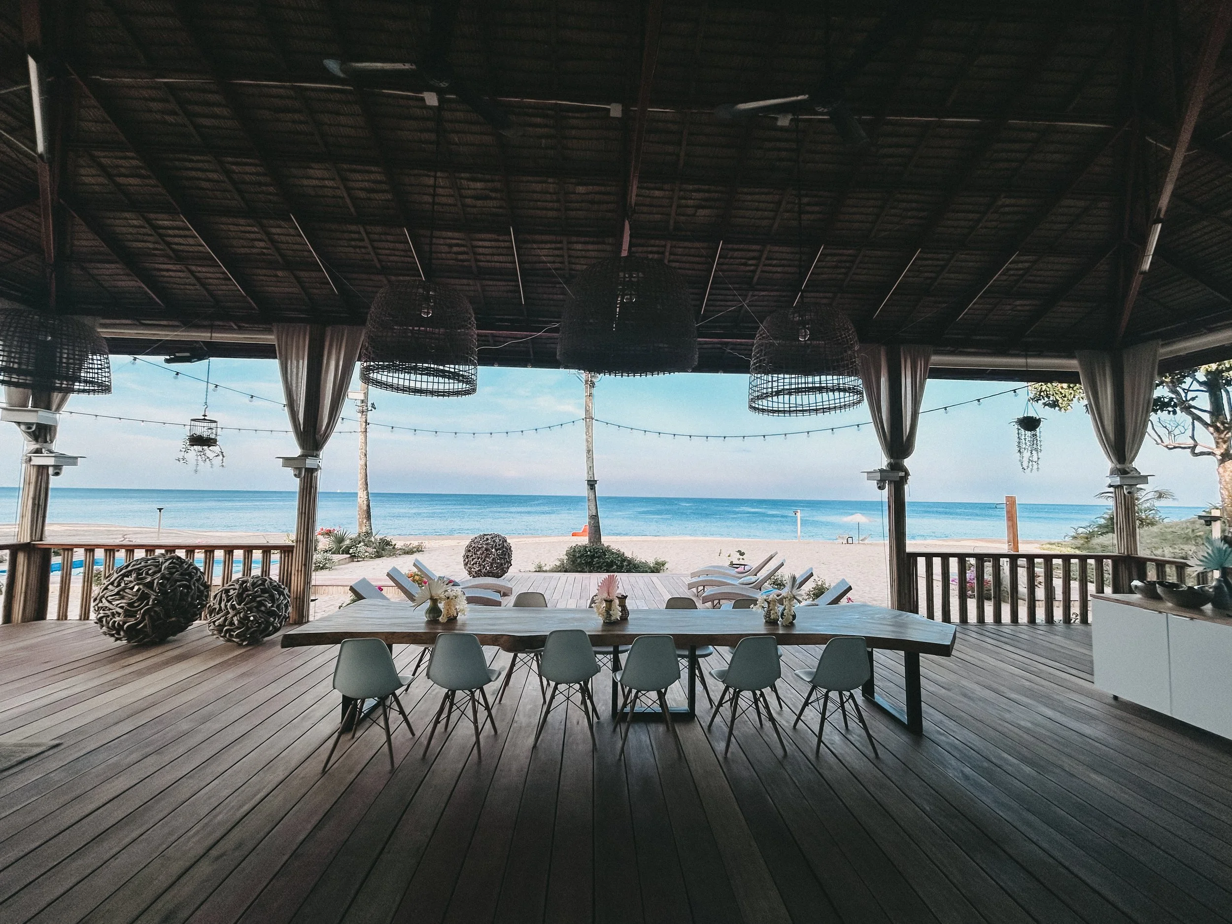 Sicht auf einen Strand mit Blick auf das Meer, gesehen durch einen offenen Pavillon mit hölzernem Boden und mehreren hängenden Korblampen.