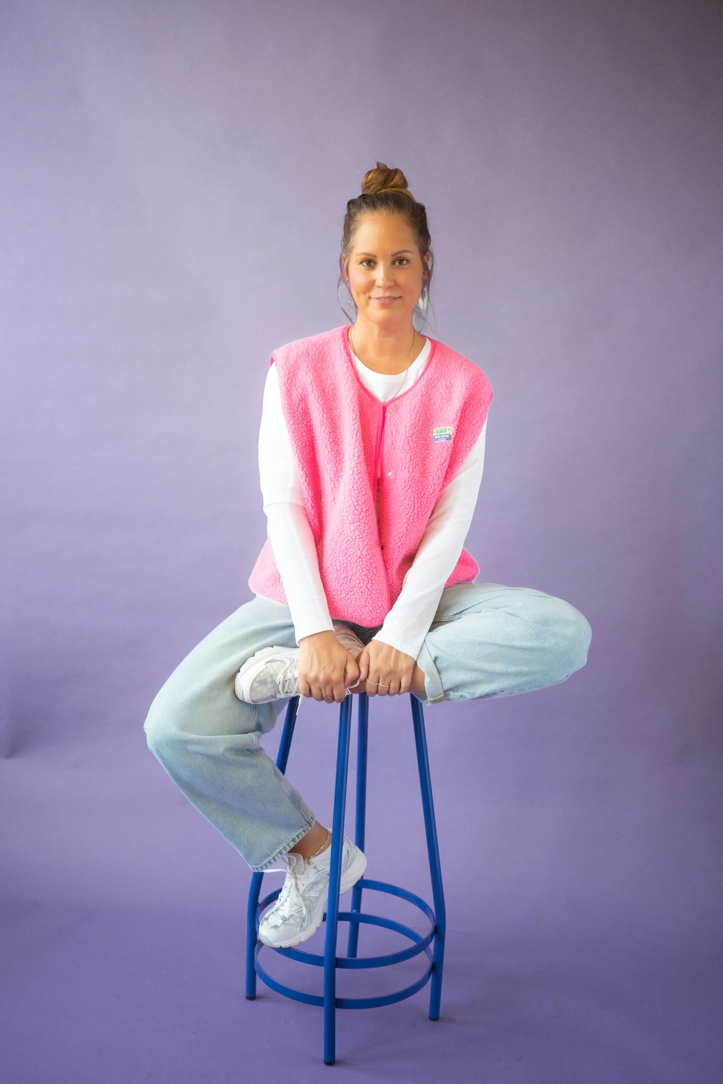 Junge Frau sitzt auf einem blauen Hocker vor einem lilafarbenen Hintergrund, trägt eine rosa Fleeceweste und sportliche Kleidung, lächelt und blickt in die Kamera.