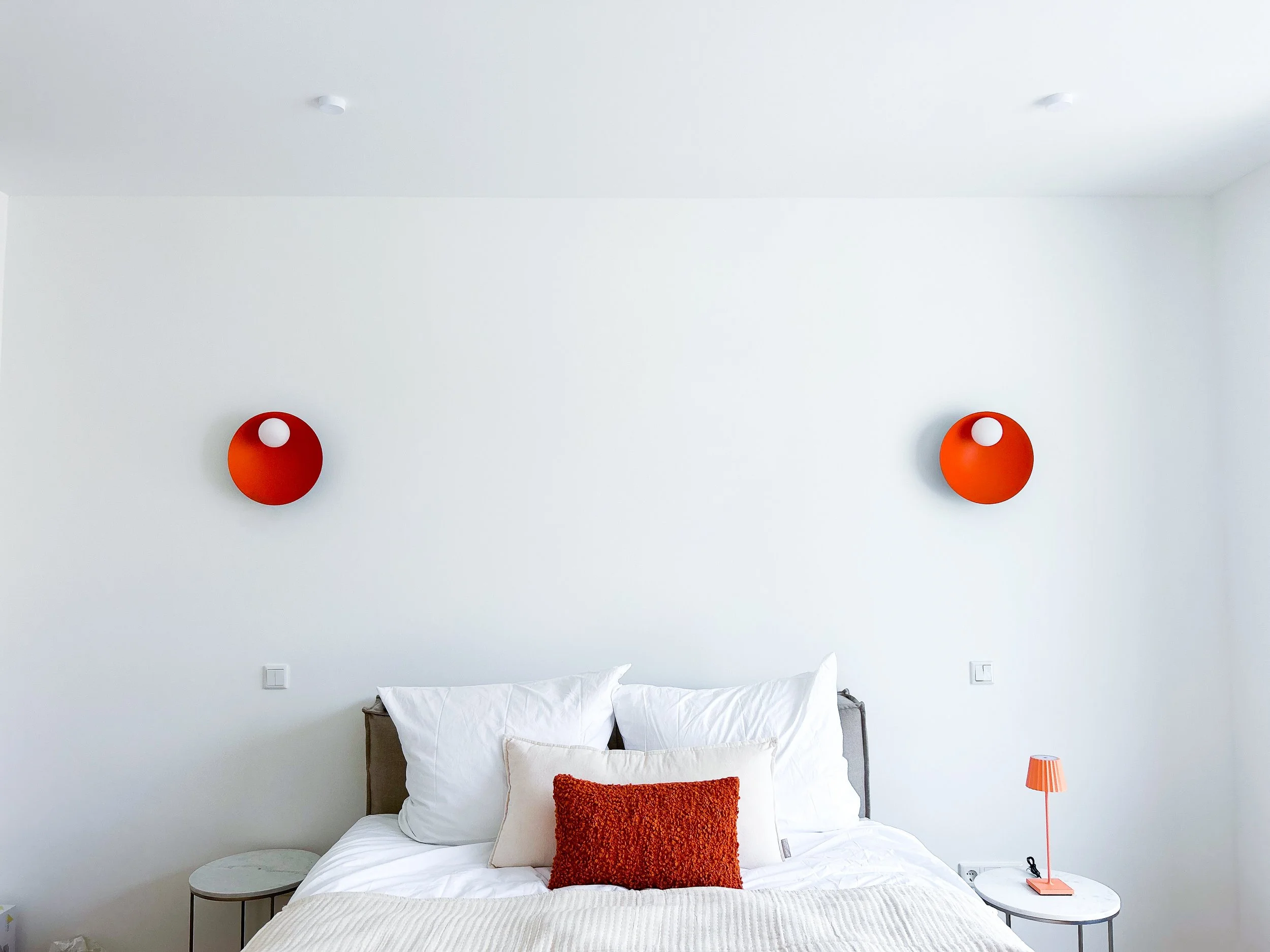 Ein modernes Schlafzimmer mit weißem Bett, zwei weißen Kissen und einem orangen Kissen. Über dem Bett hängen zwei orange Wandleuchten an der weißen Wand. Auf der rechten Seite befindet sich ein orange Nachttischlampen. Es gibt zwei weiße Stromanschlüsse an der Wand hinter dem Bett.