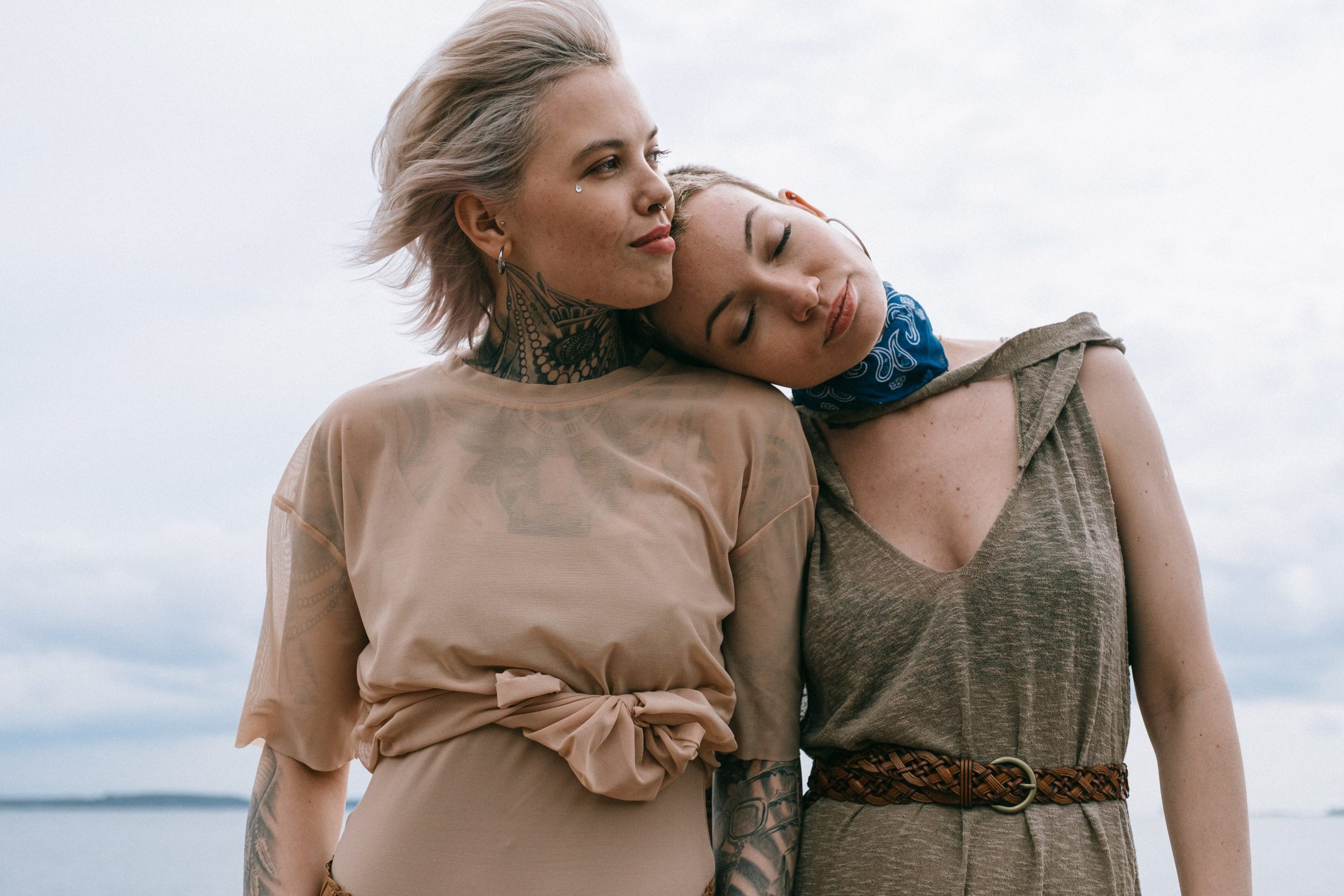 Zwei Frauen, die am Strand stehen, kuscheln und lächeln, mit einem Himmel im Hintergrund.