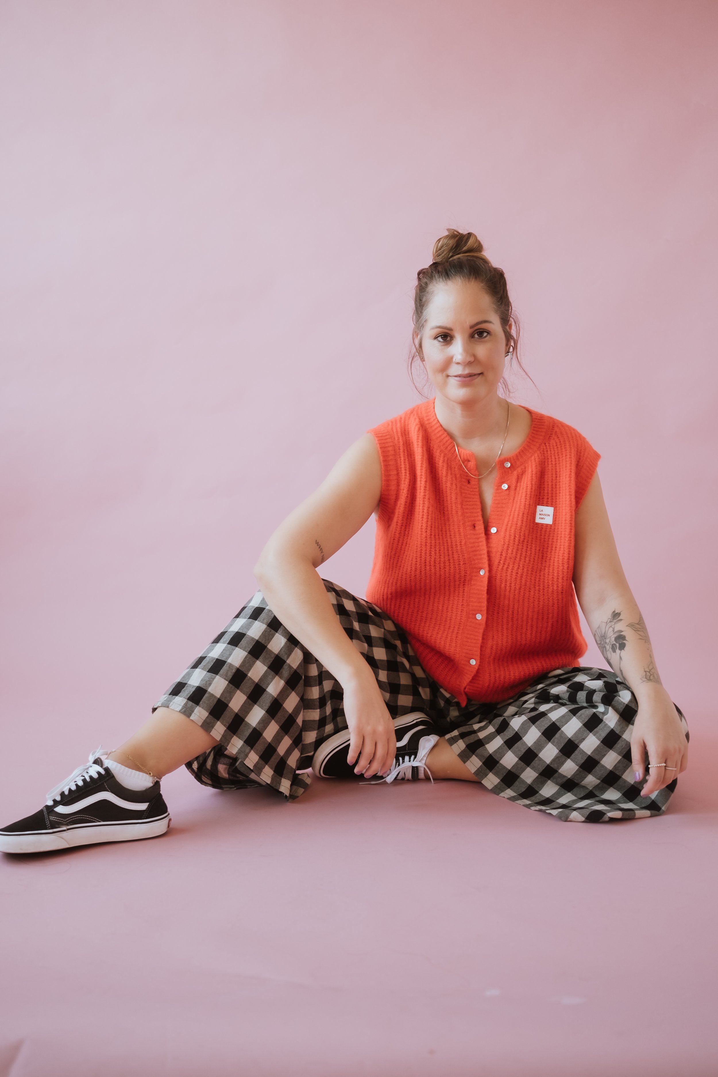 Junge Frau sitzt auf dem Boden vor rosa Hintergrund, trägt eine orangen Weste, schwarze Vans Sneakers, karierte Hosen, hat Zöpfe und Tattoos am Arm.
