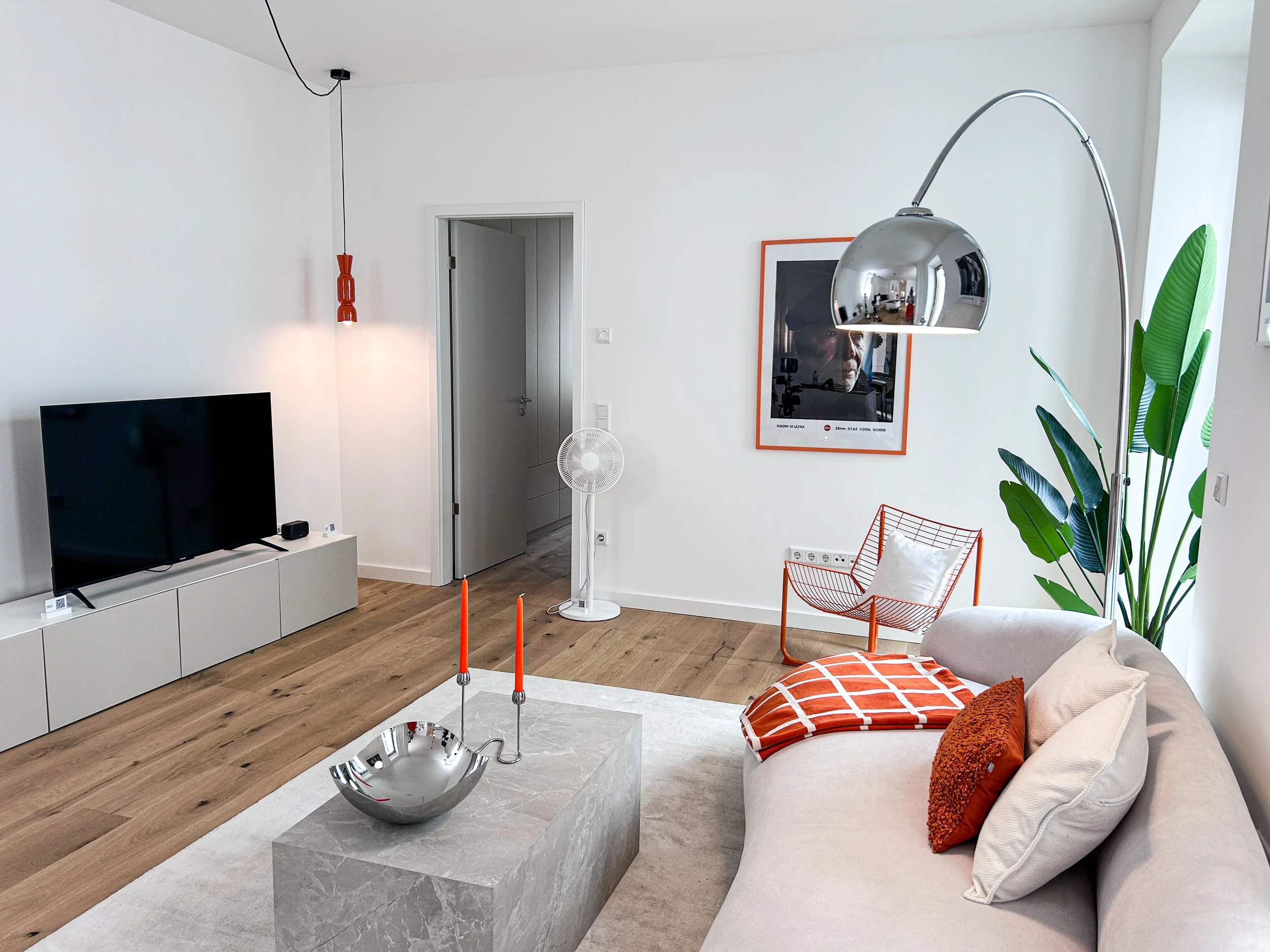 Modernes Wohnzimmer mit weißem Sofa, orange Kissen, einem schwarzen Fernseher, einem weißen TV-Schrank, einer großen grünen Zimmerpflanze, einem runden Kaffeetisch mit Kerzenhaltern, einem stilvollen Steh- oder Tischlampenschirm aus Metall, einem Spiegel mit oranger Umrandung, einer Tür im Hintergrund, und Holzboden.
