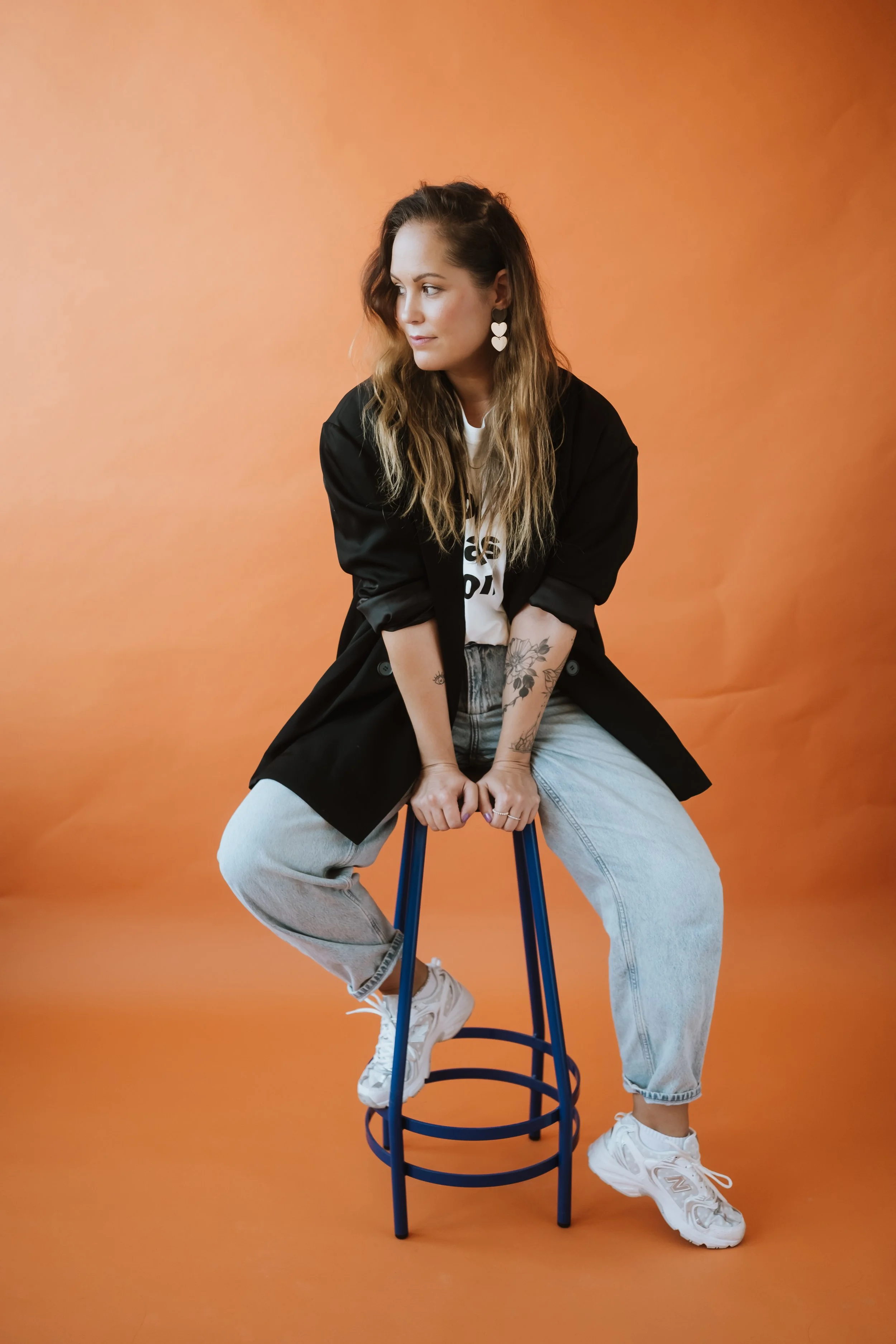 Junge Frau sitzt auf einem blauen Hocker vor einem orangefarbenen Hintergrund, trägt weiße Turnschuhe, Jeans, ein weißes T-Shirt und schwarze Jacke, hat lange, gewellte Haare, große Herzohrringe und Tattoo an den Armen, blickt nach links.