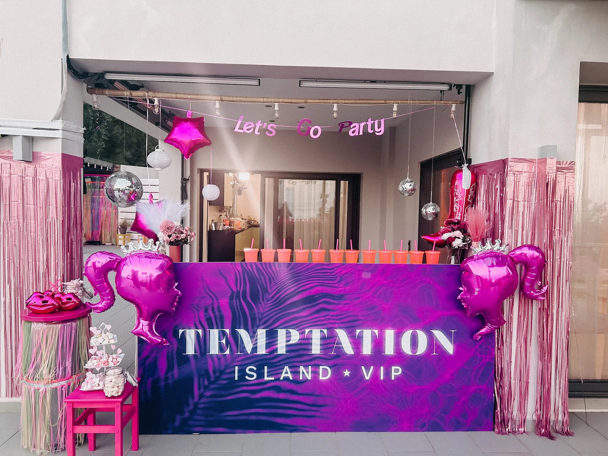Dekoration für eine Party mit pinkfarbenen Ballons, Papierlampen, einem pinken Schreibtisch, einem Banner mit der Aufschrift 'Let's Go Party', einem Drinkestand mit Strohhalmen, Pinkelavy, Blumen und einem pinken Vorhang. Der Hintergrund zeigt den Eingang zu einem Gebäude.