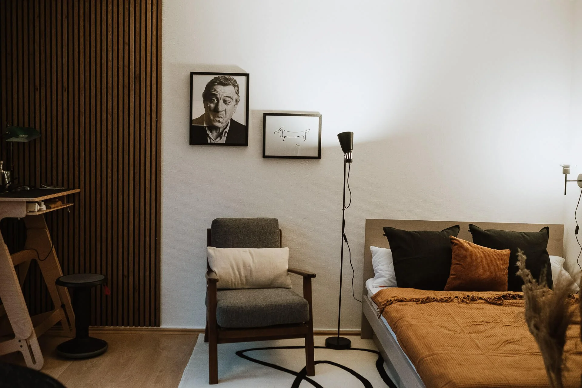 Ein modernes Schlafzimmer mit einer weißen Wand, schwarzen Kissen, brauner Decke, einem Bett, einem Stuhl, einem Bilderrahmen mit Portrait und einer Zeichnung eines Dackels, einem Stehlampe, und einem offenen Kleiderschrank aus Holz.