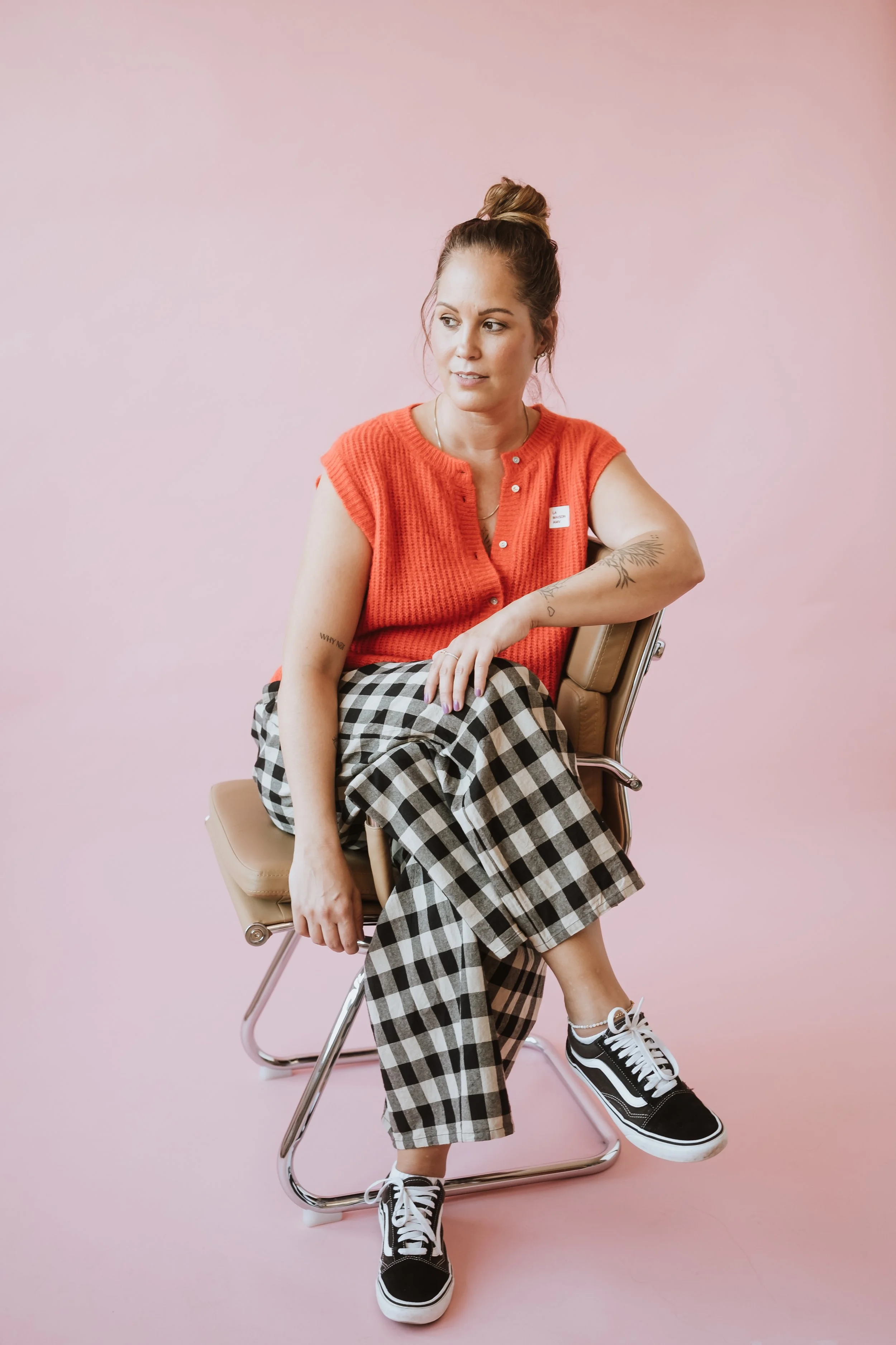 Junge Frau mit buntem Look, sitzend auf einem Stuhl vor pinkem Hintergrund, trägt schwarz-weiße Vans Sneaker, kariertes Hosenpaar und orangene Weste, hat Tattoos am Arm, Haar im Dutt, schaut seitlich.