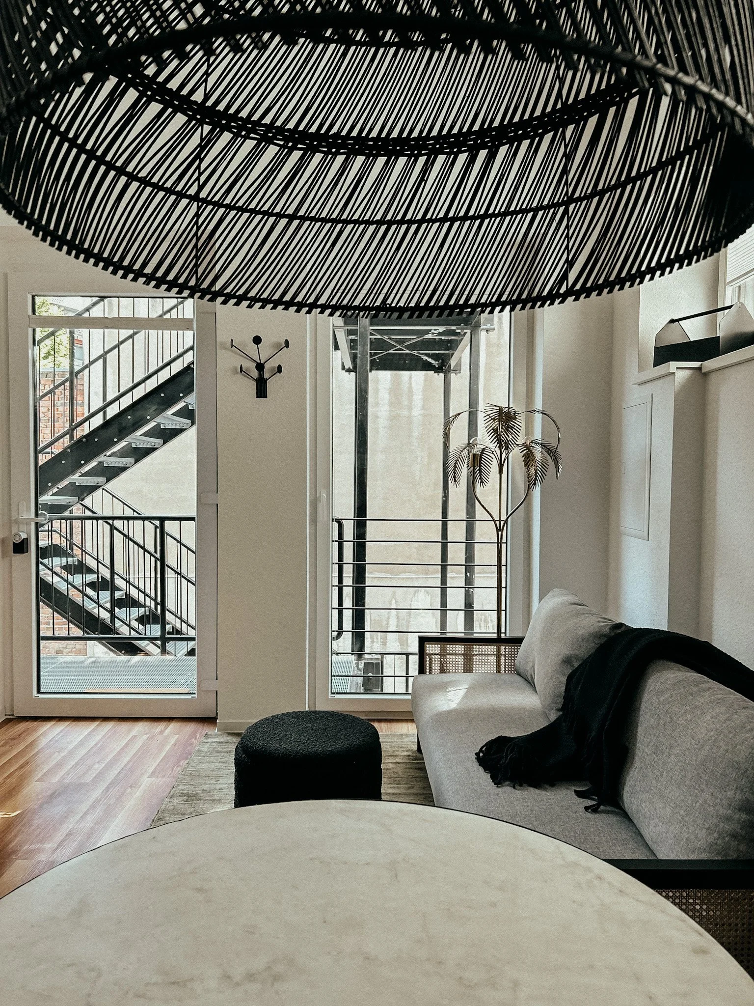 Wohnzimmer mit grauem Sofa, direkter Blick auf Tonpappentisch, schwarzem Hocker, auf dem Sofa liegt schwarzer Schal. Große Fenster, glatte weiße Wände, Stehleuchte und Kletterpflanze. Treppe im Hintergrund, Balkon mit Geländer, indirekte Beleuchtung.
