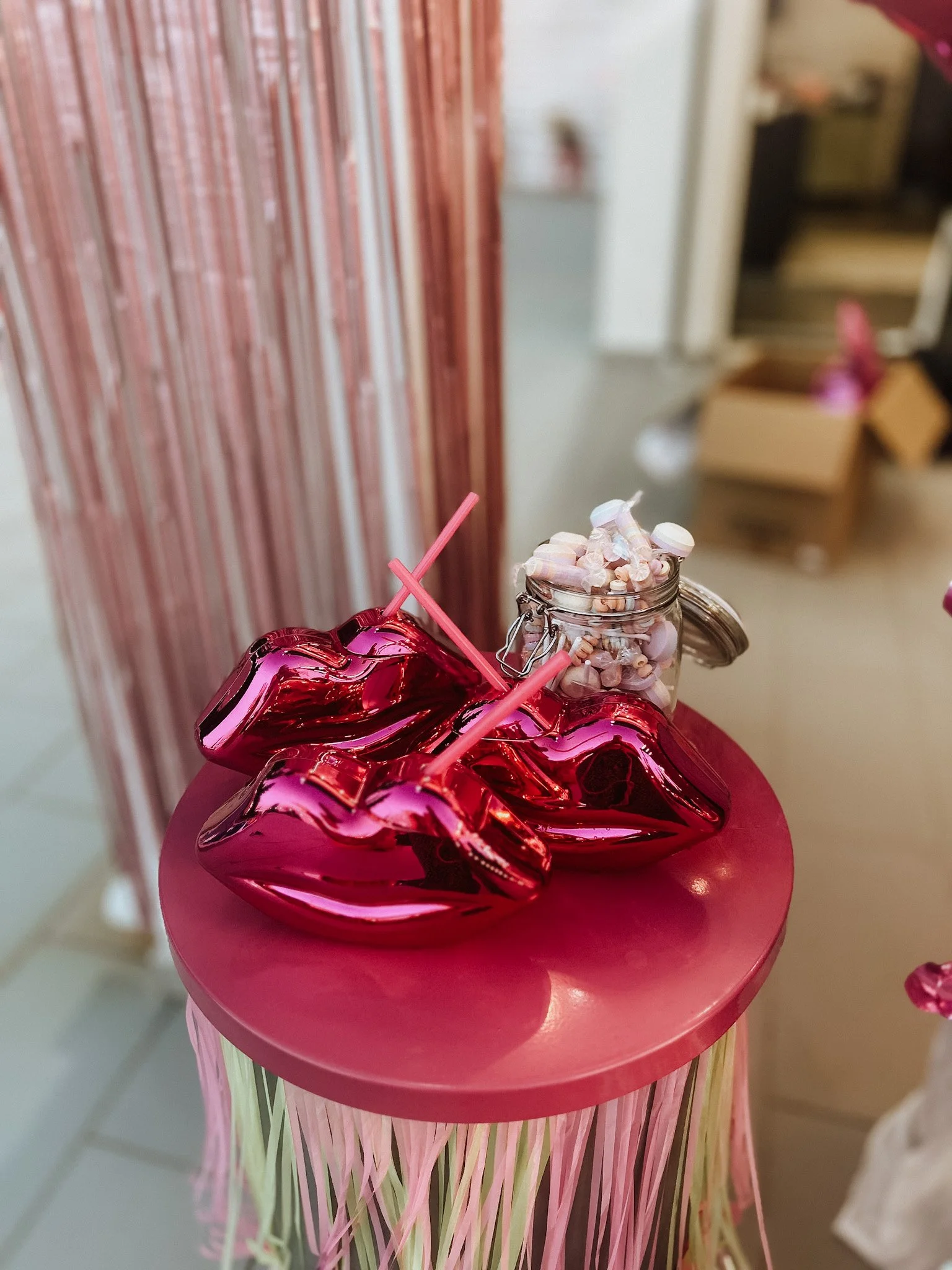 Ein dekorativer Tisch mit einer pinkfarbenen Tischdecke und mehreren Herzballons. Auf dem Tisch steht ein Glas mit Süßigkeiten und Trinkhalmen, im Hintergrund sind regale und andere Raumdetails sichtbar.