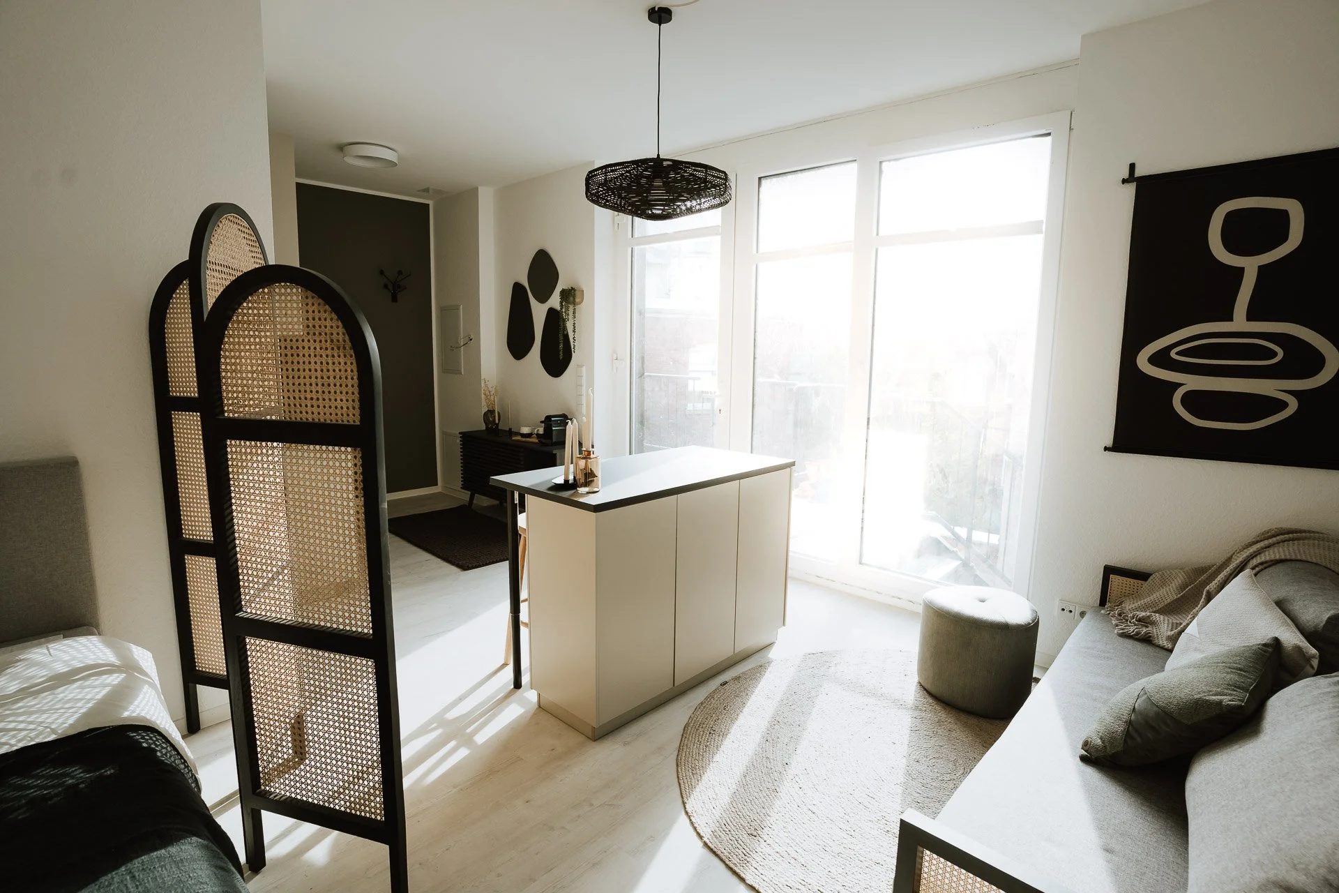 Heller moderner Wohnraum mit großem Fenster, Sofa, kleiner Fußhocker und Trennwand aus Rattan. Deko mit schwarzen Wandobjekten und Pflanzen, gemütliches Ambiente mit natürlichem Licht.