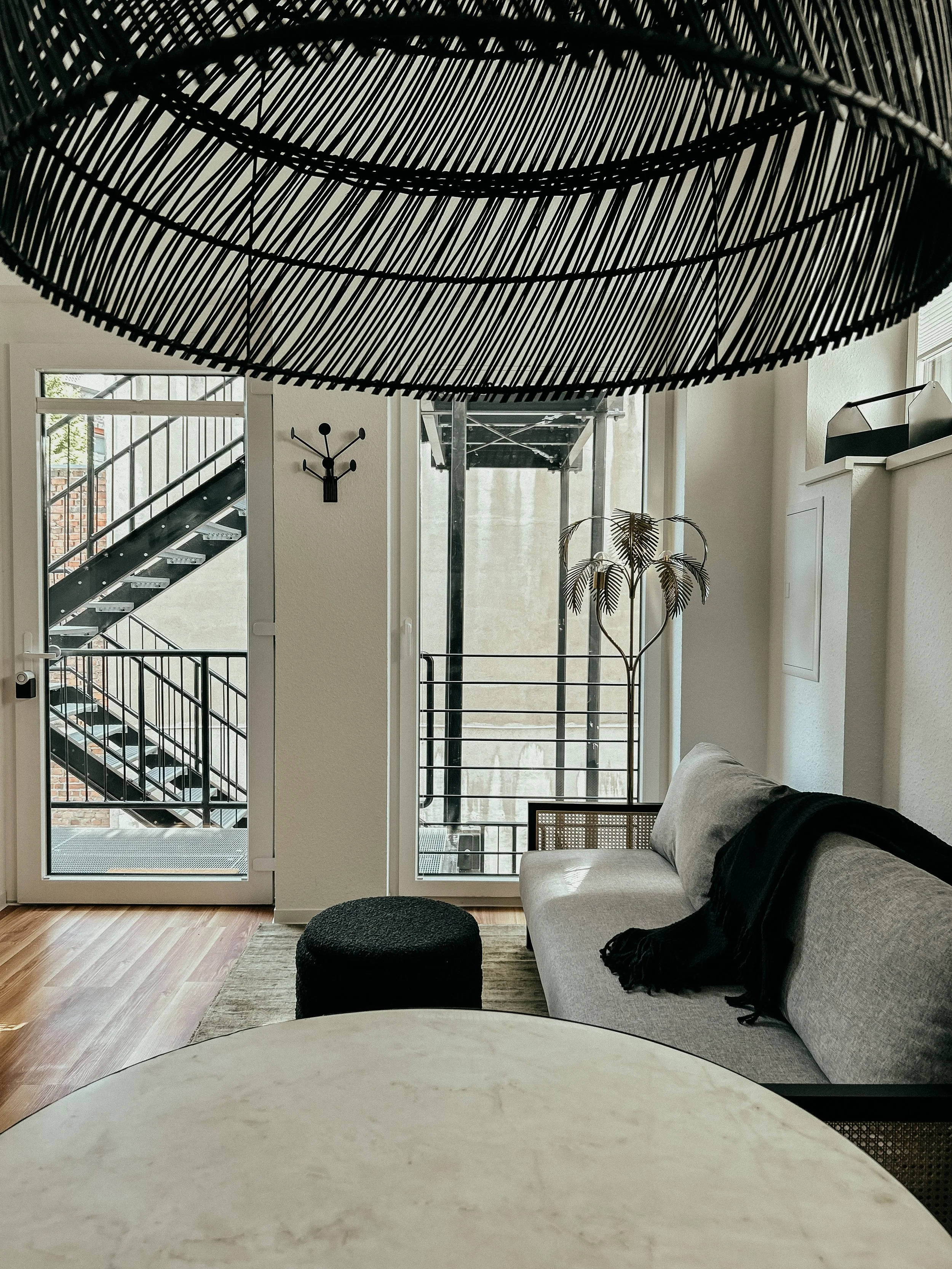 Innenraum eines Wohnzimmers mit grauem Sofa, schwarzen Kissen, einem schwarzen Hocker, einem grauen Teppich, großen Fenstern, einer Tür mit Glas und einer modernen Wanddekoration, einer Pflanze und einer Treppe im Hintergrund.