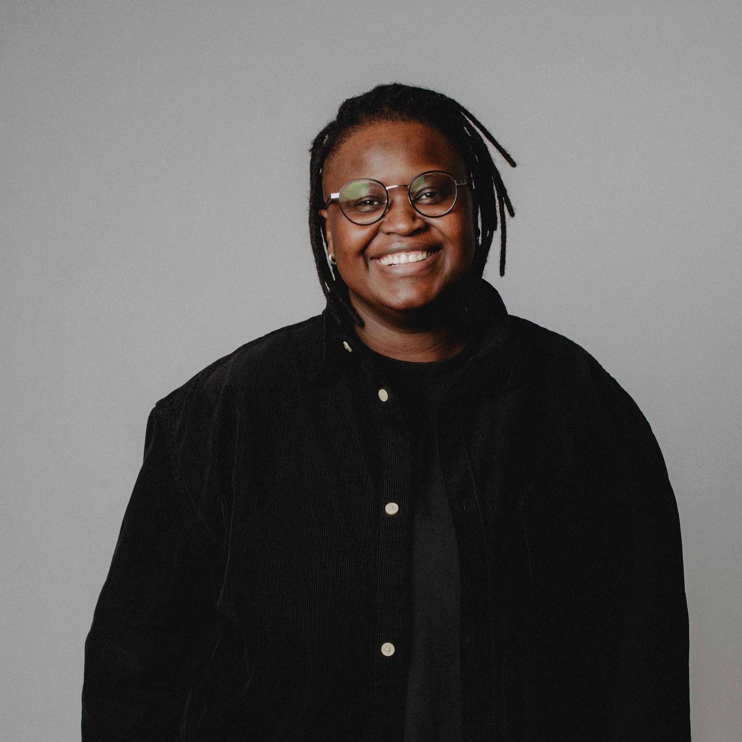 A woman with glasses and dreadlocks smiling at the camera, wearing a black jacket against a plain gray background.