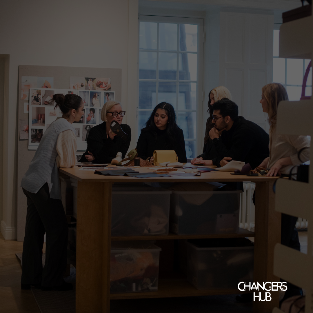 People attending a discussion or seminar, seated in chairs indoors near large windows, with some taking notes and one person speaking.