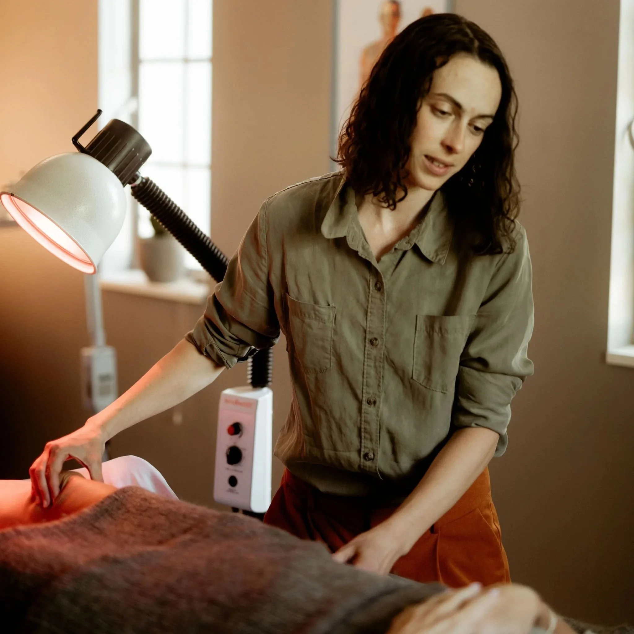 A woman with dark curly hair wearing an olive-green shirt, sitting at a table in a room with natural light, working or reading with a lamp nearby.