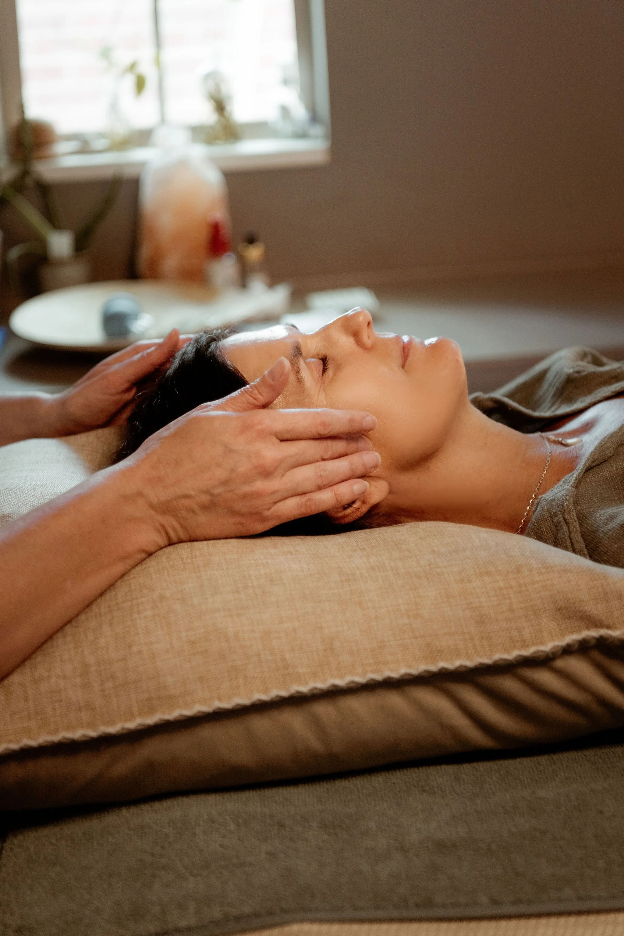 A woman lying down on a massage table, receiving a facial treatment. A person is gently touching her face, possibly applying a skincare product or performing a massage. The setting appears to be a spa or skincare clinic.