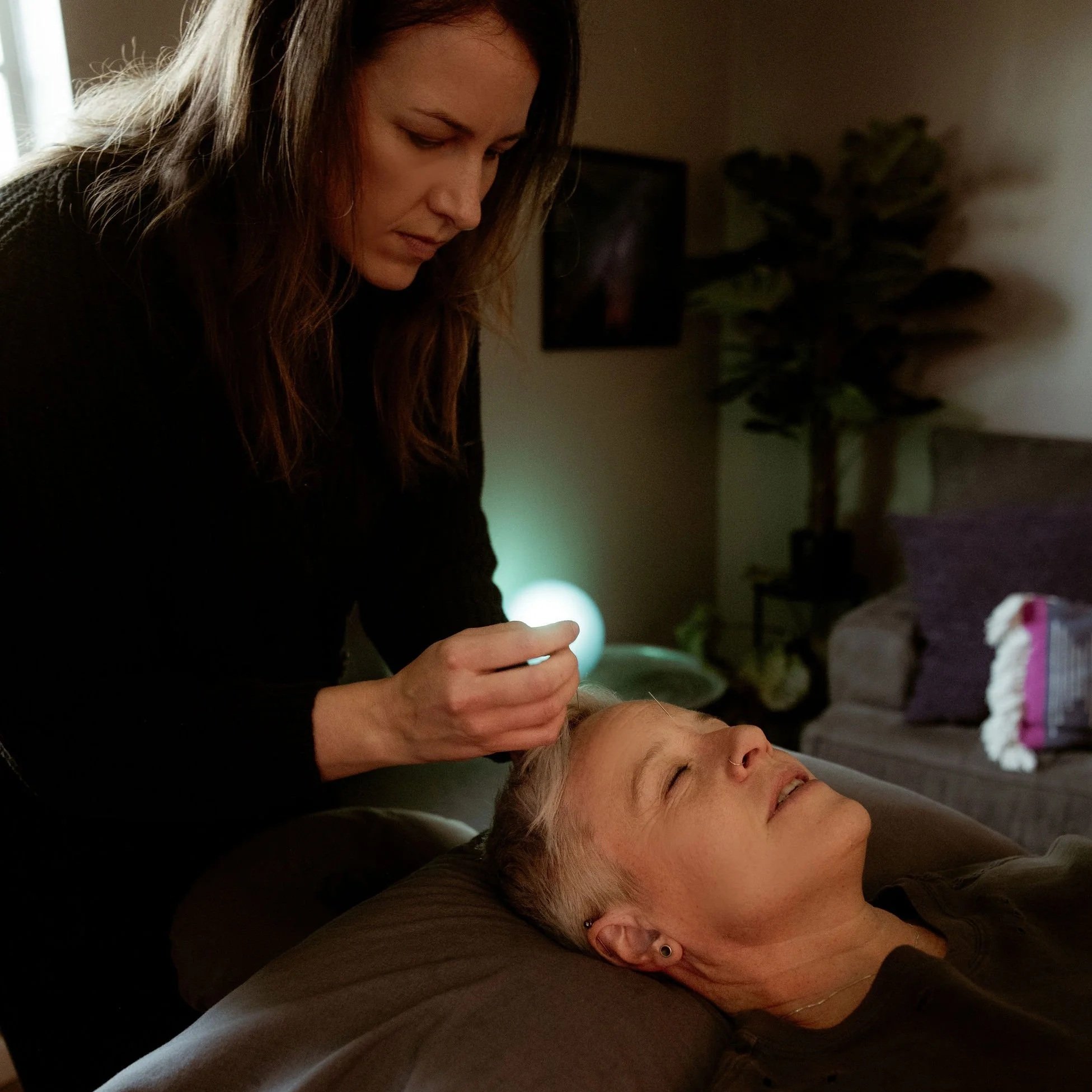 A woman with long brown hair receiving a head massage from another woman in a dimly lit room with a houseplant and TV in the background.