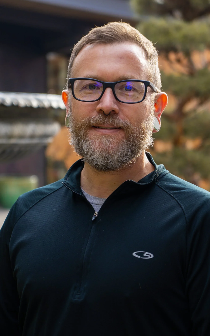 A light-skinned man with glasses and a beard smiling outdoors, wearing a black zip-up jacket.