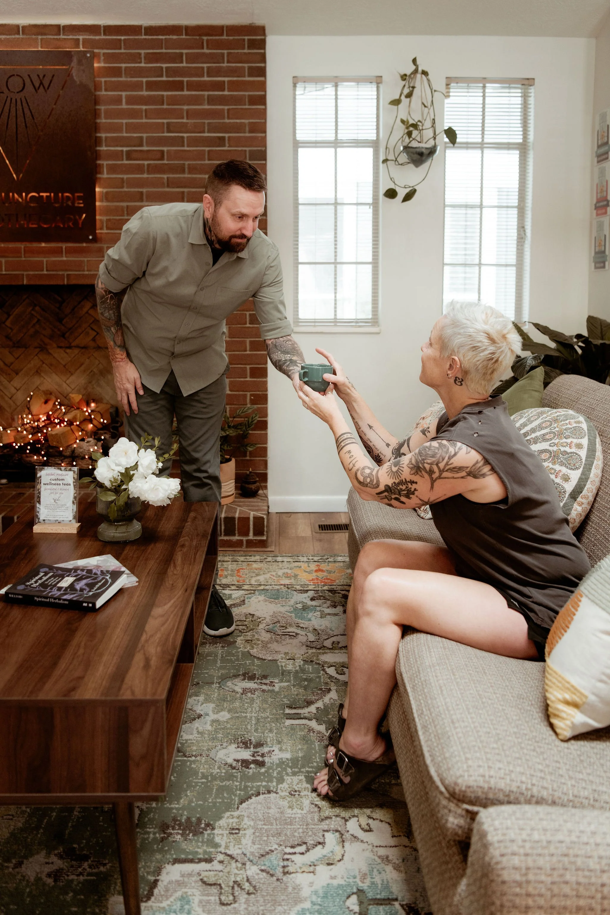 A man with tattoos on his arms handing a green mug to an older woman with short white hair sitting on a beige couch in a cozy living room. They are near a brick fireplace decorated with string lights and flowers on a table. The room has large windows, hanging plants, and framed pictures on the wall.