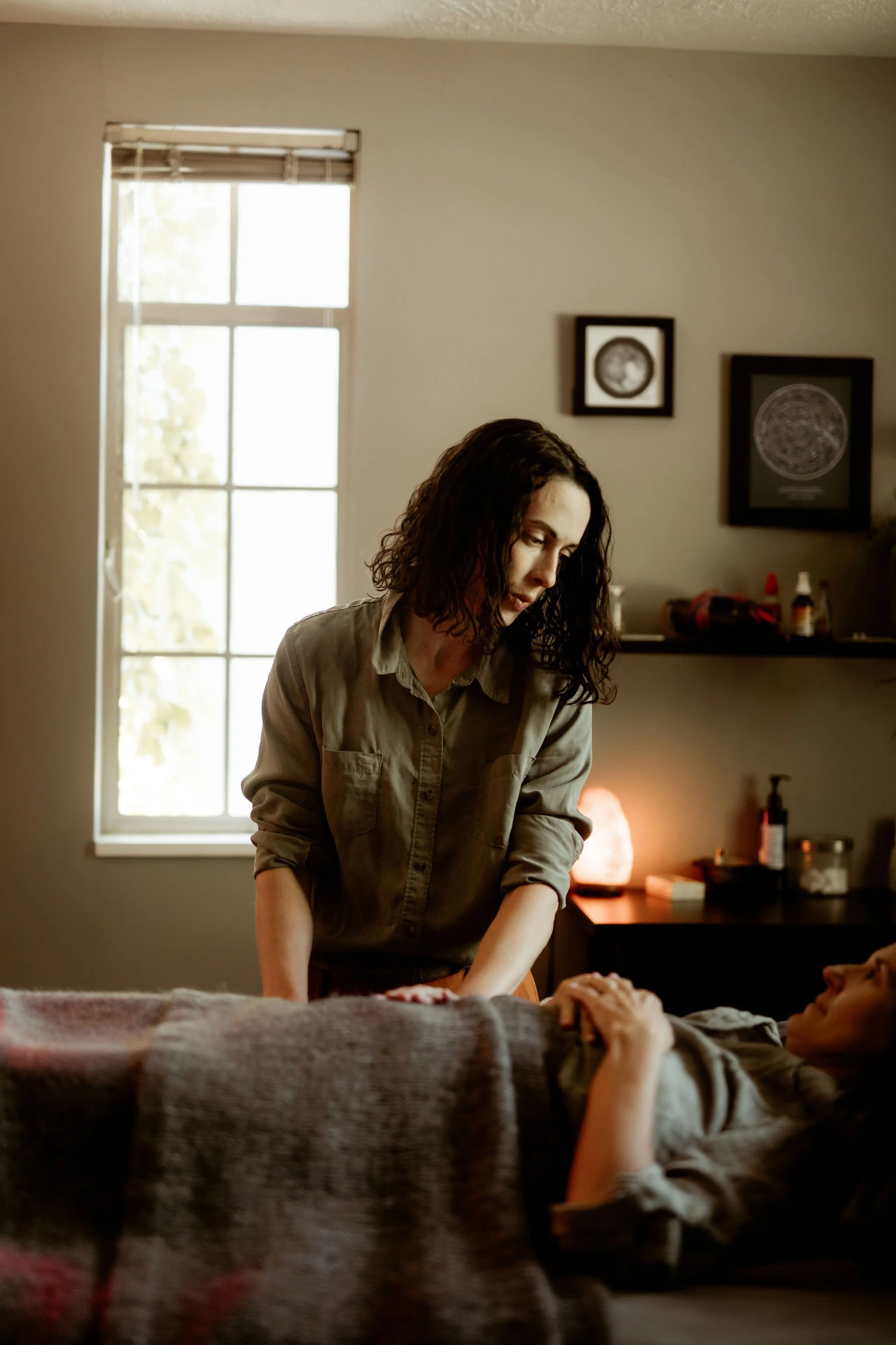 A woman with dark curly hair wearing a beige button-up shirt leans over a person lying on a couch. The person on the couch is holding the woman's hand. The scene is set in a room with natural light coming through a window, framed pictures on the wall, and various items on a shelf in the background.