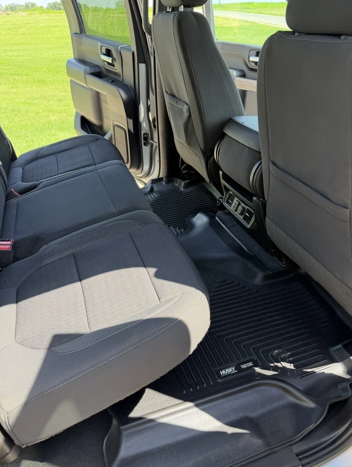 The interior of a pickup truck showing the back seat area with fabric seats, black rubber floor mats with the Husky brand, and the back of the front seats, with a view of the green field outside through the open door.
