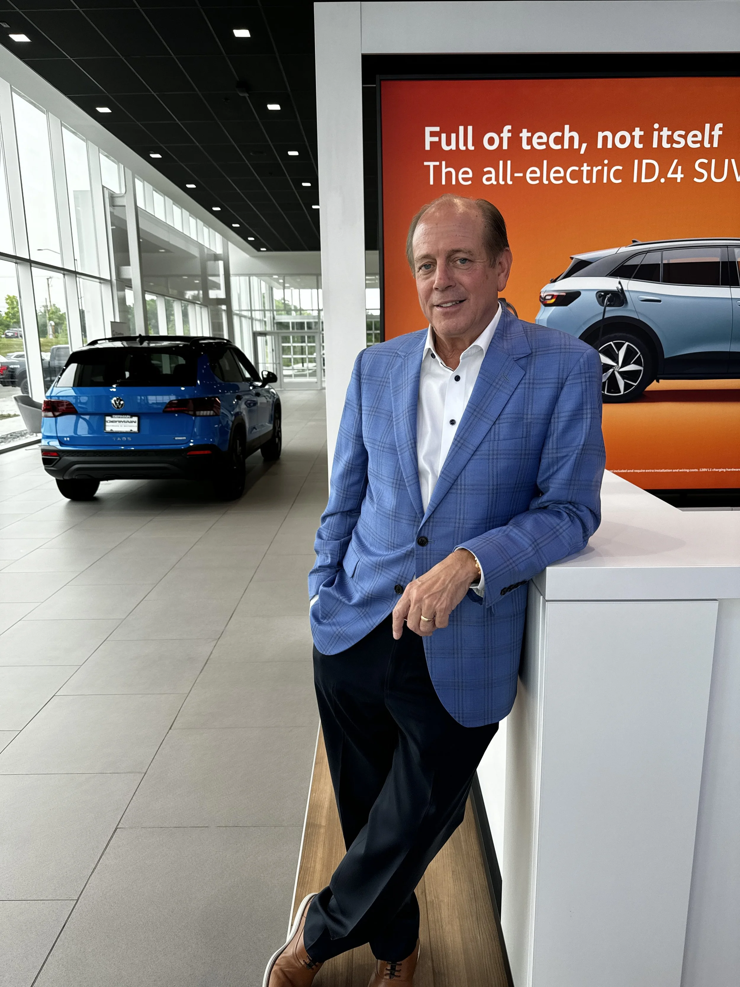 A man in a blue plaid blazer and white shirt standing inside a car dealership, with a blue SUV behind him and a large orange advertisement for a full electric SUV.
