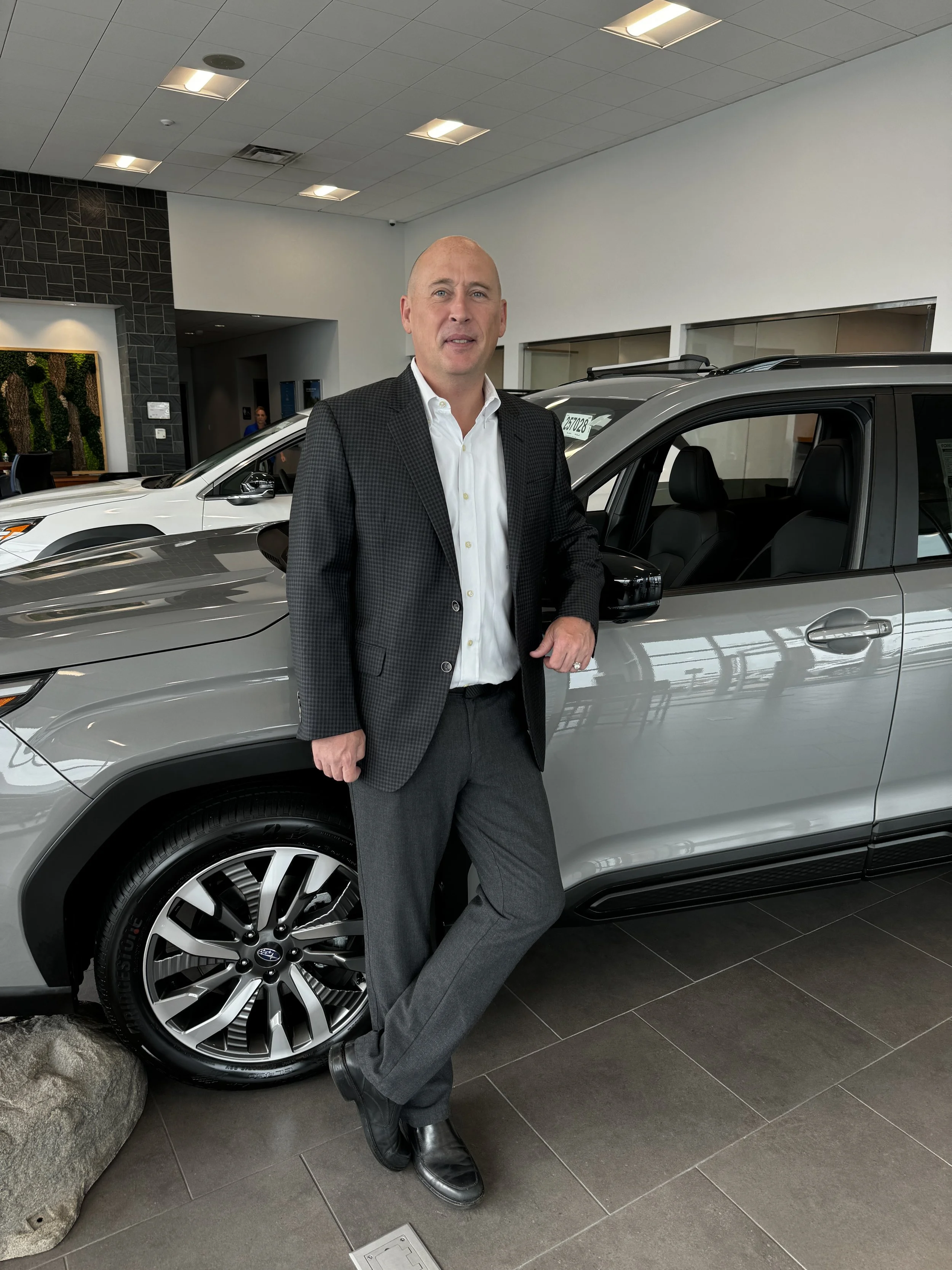 A man in a suit standing inside a car dealership, leaning against a gray car with a black interior, with other cars visible in the background.