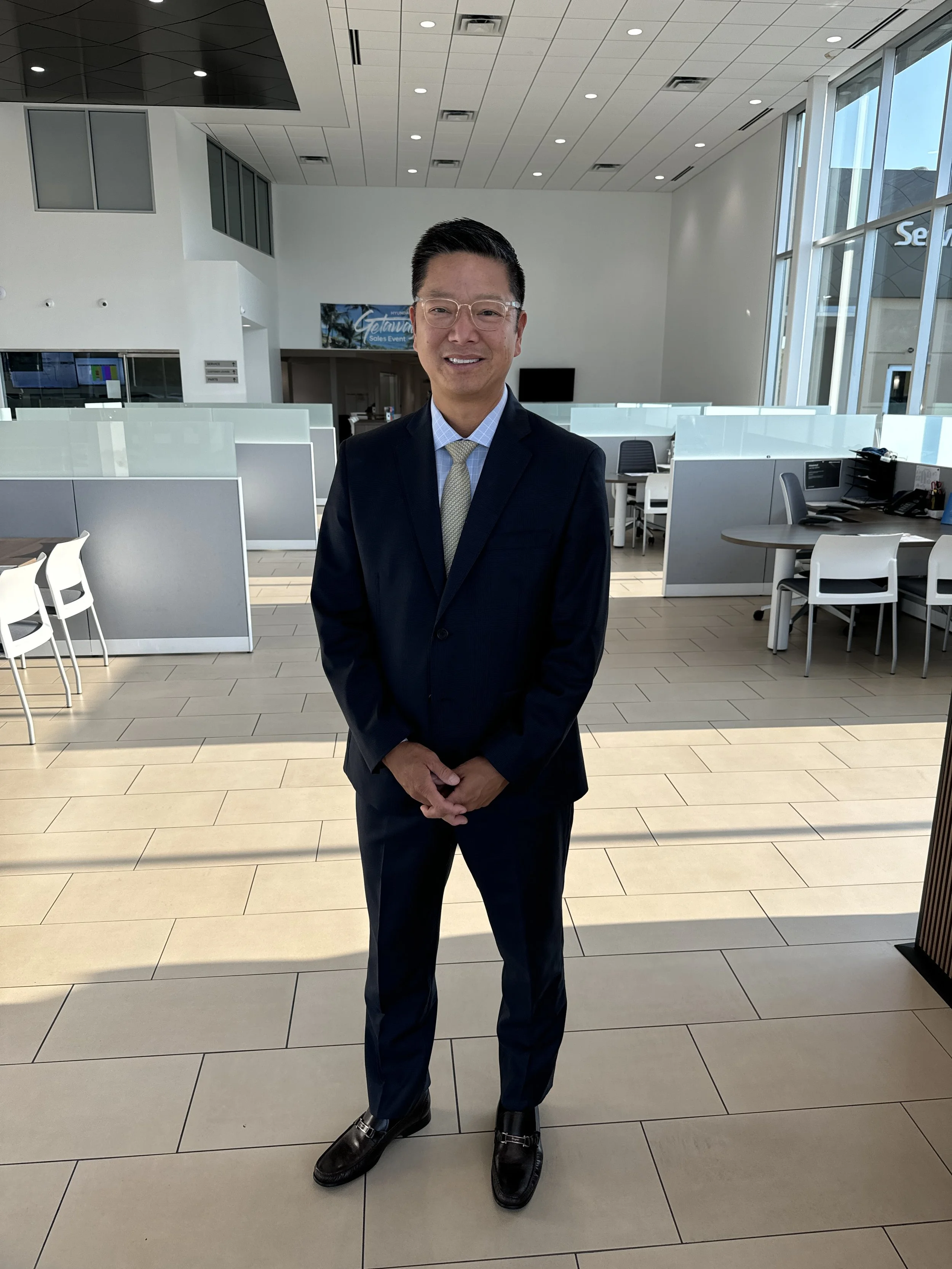 A man in a dark suit and tie standing in a modern office lobby with large windows and white partitioned workspaces.