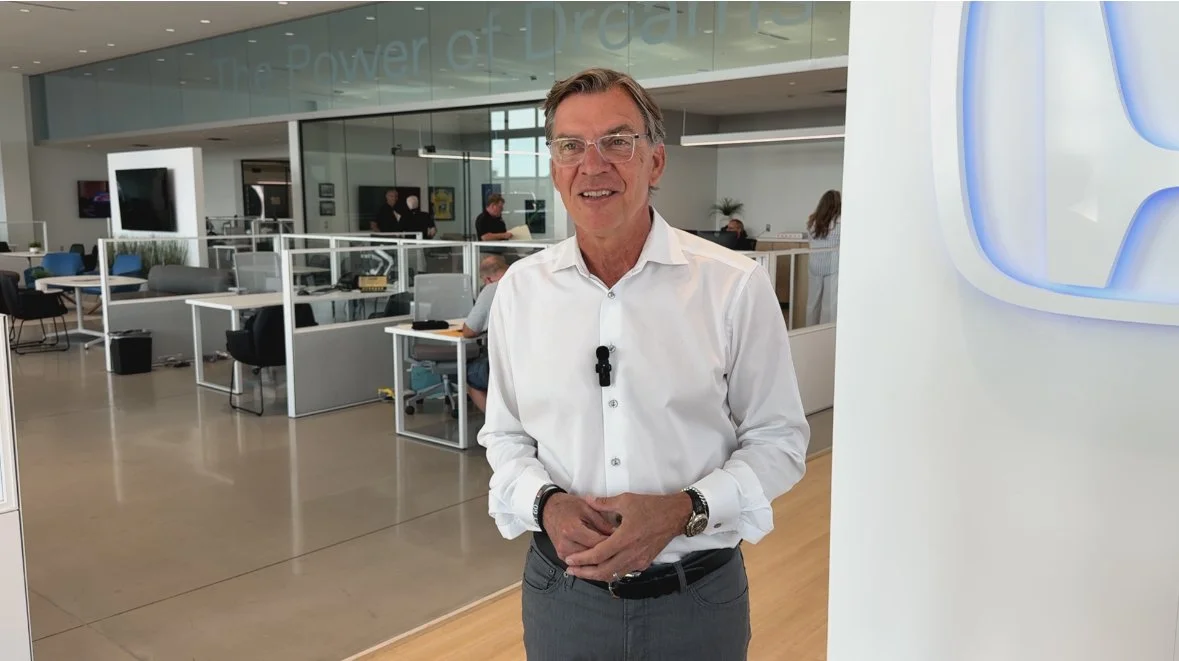 A man wearing glasses and a white shirt stands inside a modern office space with glass walls, desks, and people working in the background.