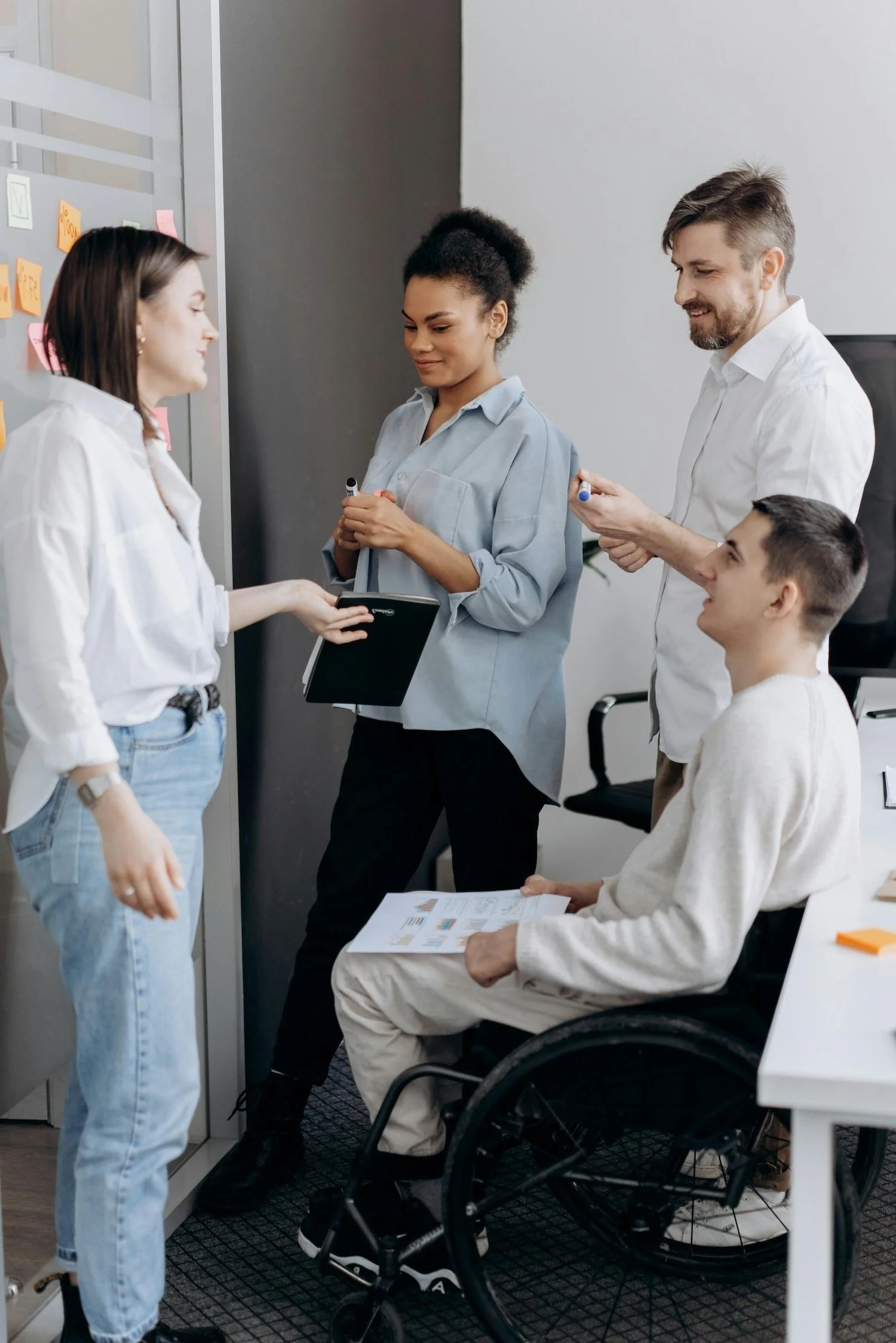 Four people having a discussion in an office, one person using a wheelchair, holding paper documents and another person holding a notebook.