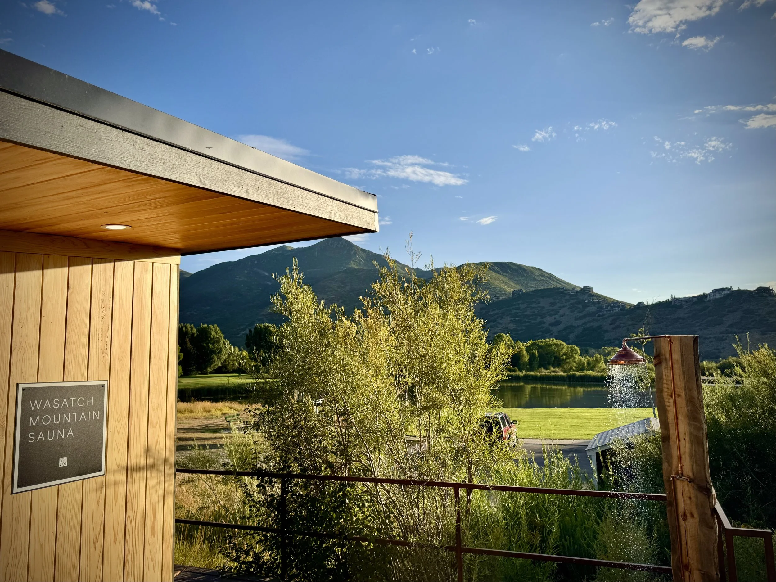 Outdoor scene with a wooden building labeled 'Wasatch Mountain Sauna' on the left, lush green trees, a lake, and mountains in the background under a partly cloudy blue sky.