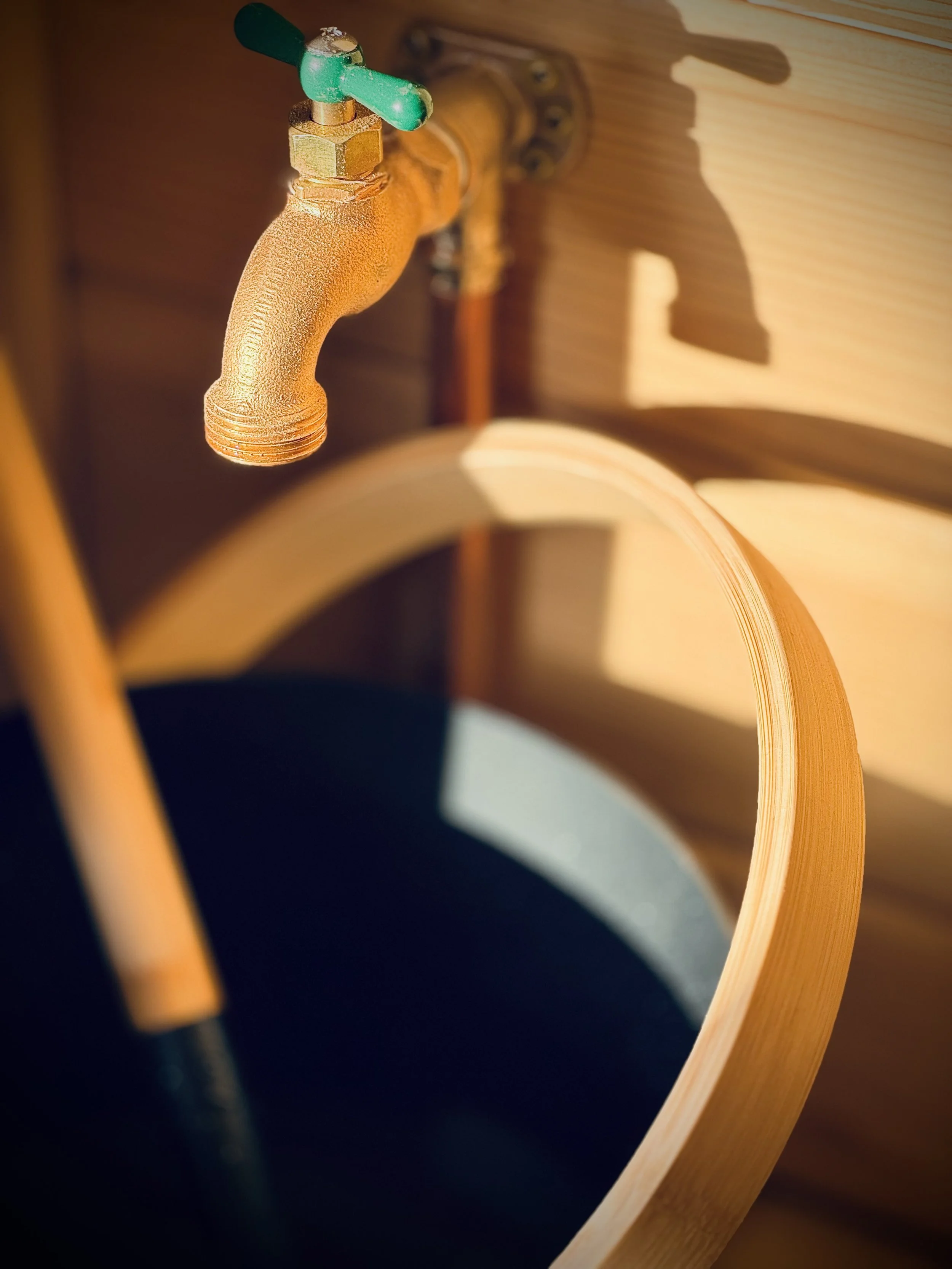 Close-up of an outdoor faucet with a green handle, mounted on a wooden wall, casting a shadow. A wooden bucket is partially visible beneath it.