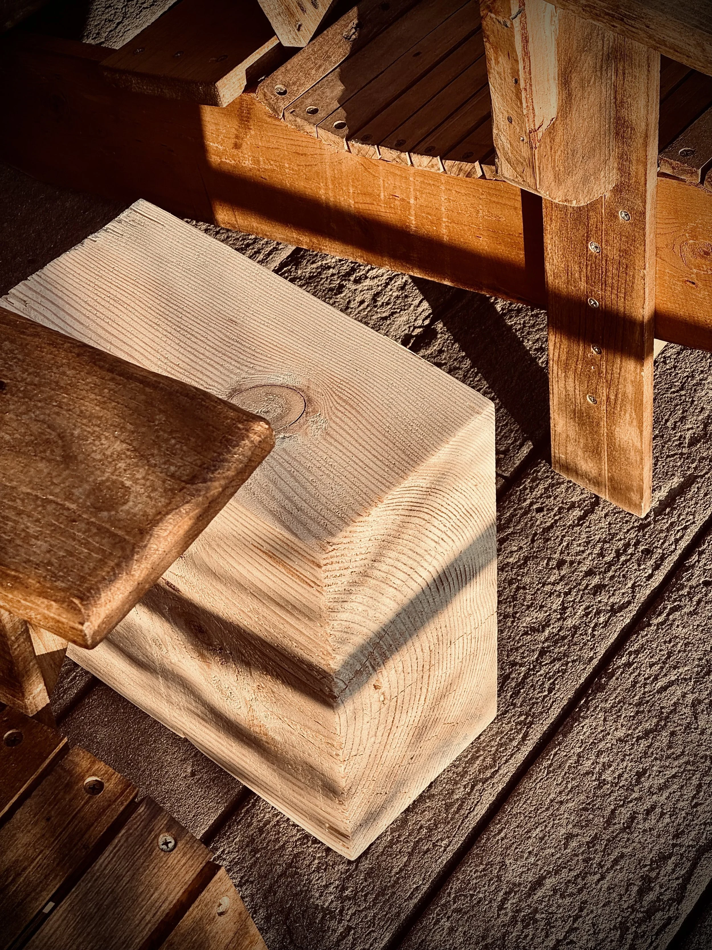 Close-up of wooden furniture pieces, including a light-colored wooden block, a darker wooden plank, and part of a wooden chair or table, with shadows cast on a rough concrete surface.