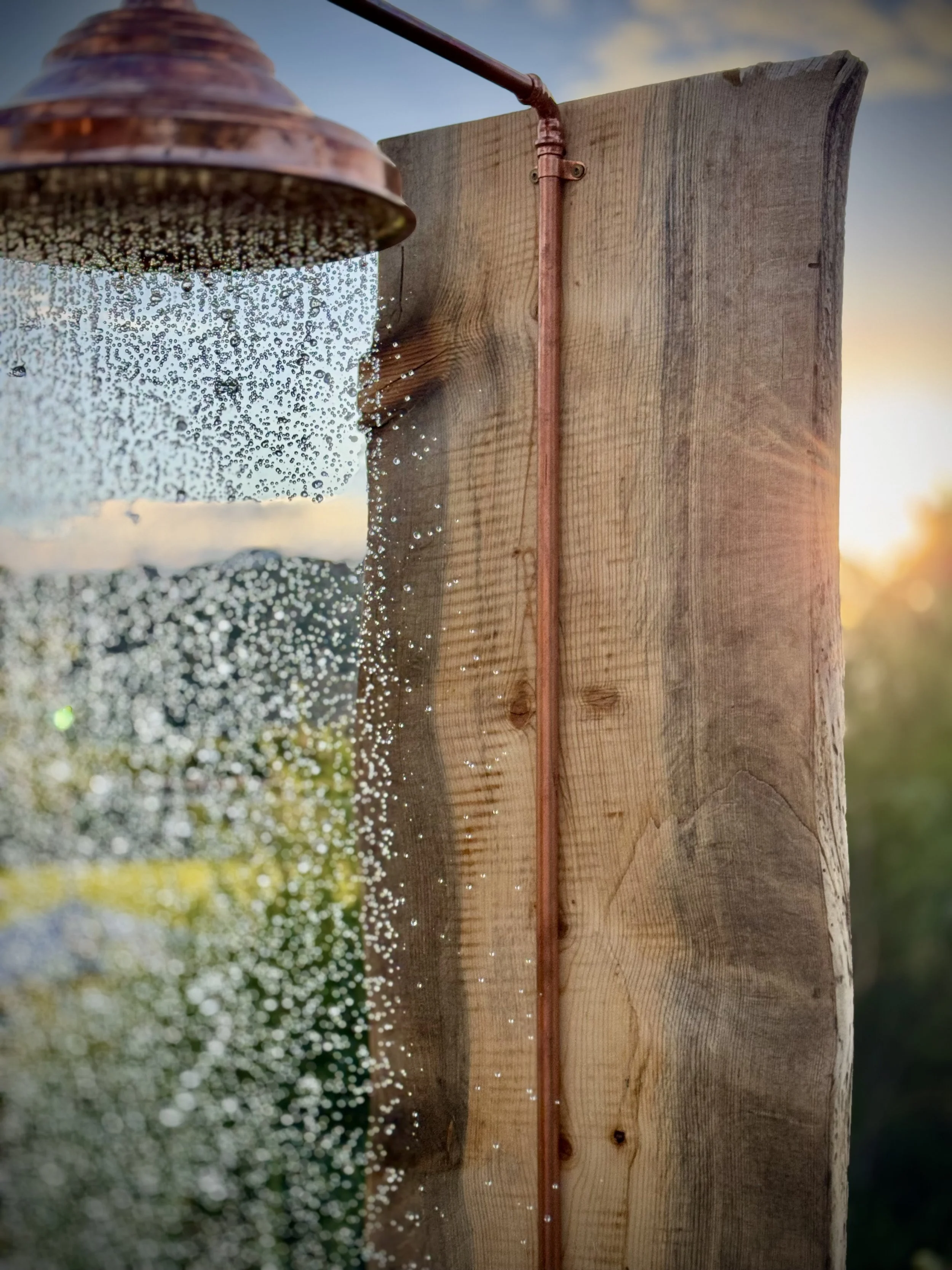Close-up of an outdoor shower with a copper pipe, a showerhead, and a wooden post, with water droplets and a sunset in the background.