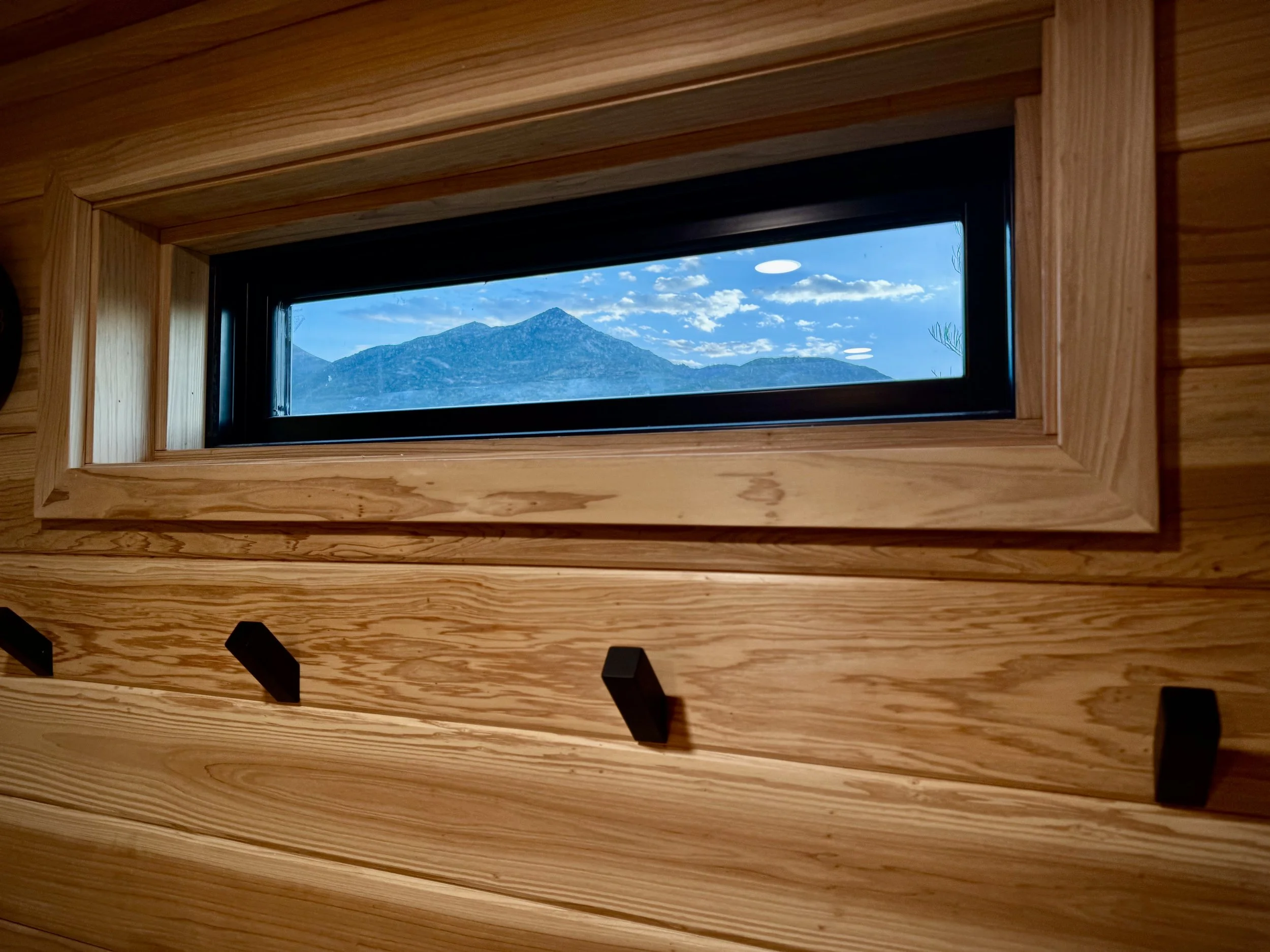 A horizontal rectangular window with a black frame, set in a wooden wall, showing a mountain range under a partly cloudy sky.