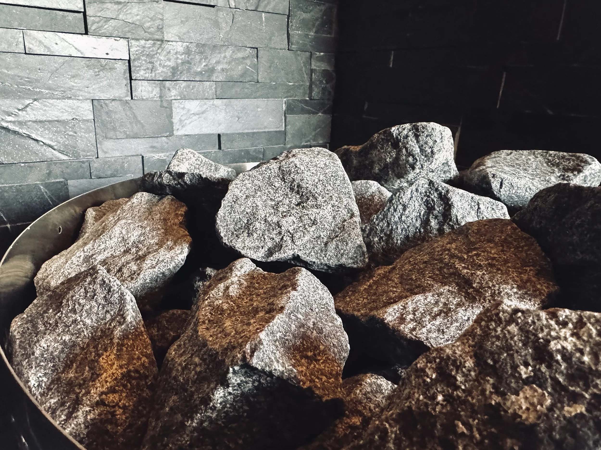 Close-up of a pile of large, irregularly shaped rocks or stones in a metal container in front of a dark stone wall.