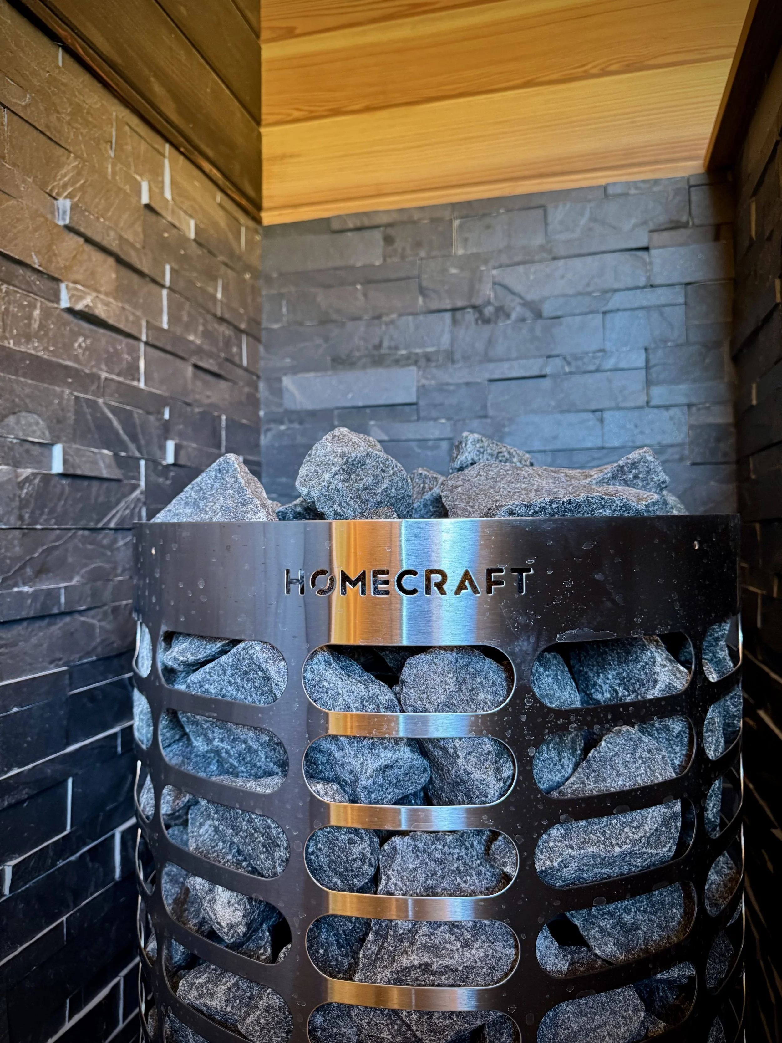 A modern sauna heater filled with rocks, with the brand name 'Homecraft' engraved on the metal band around it, set against dark stone brick walls.