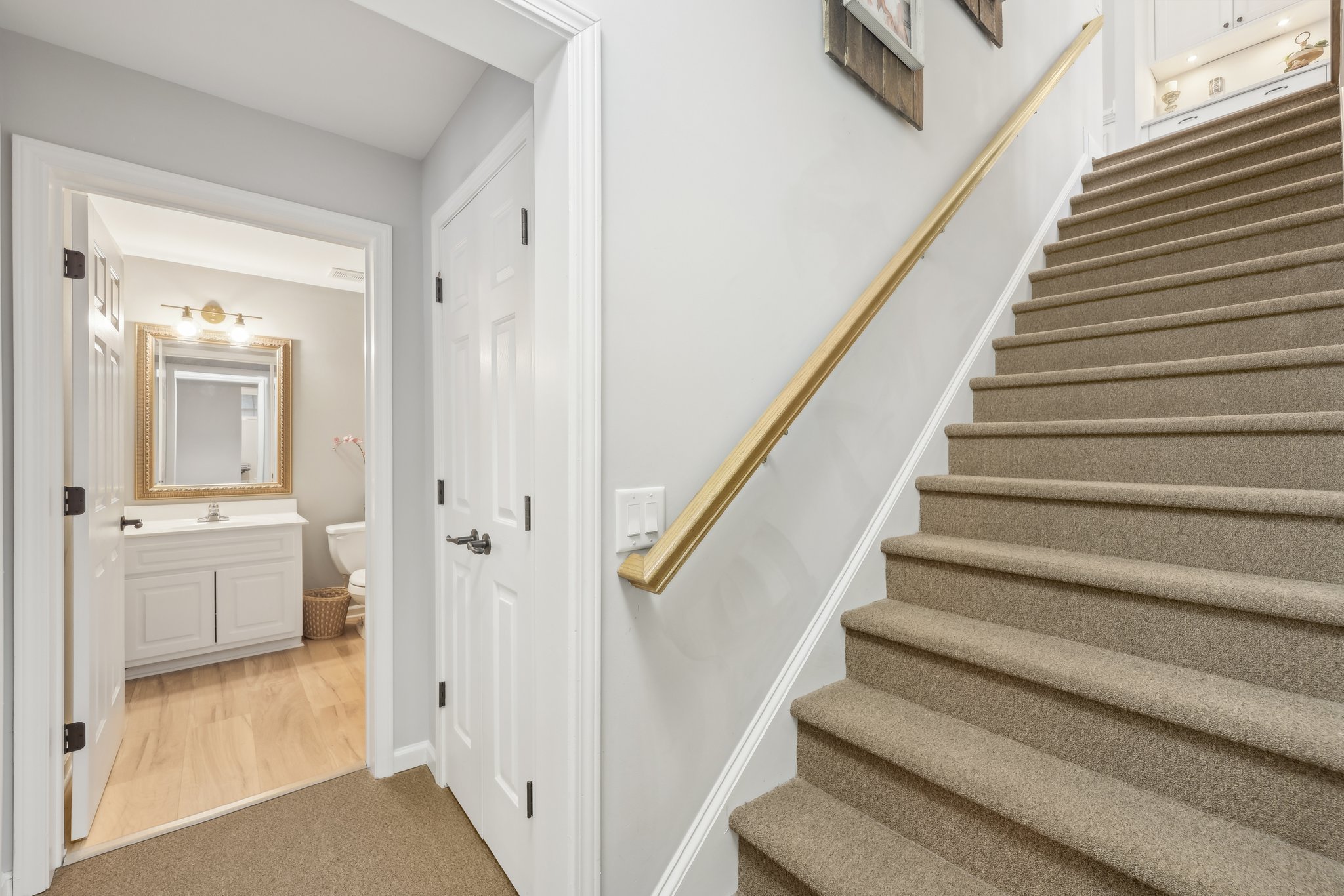 Interior of a house featuring a carpeted staircase with a wooden handrail, a hallway with a closed door, and an open door leading to a bathroom with a vanity, mirror, and toilet.