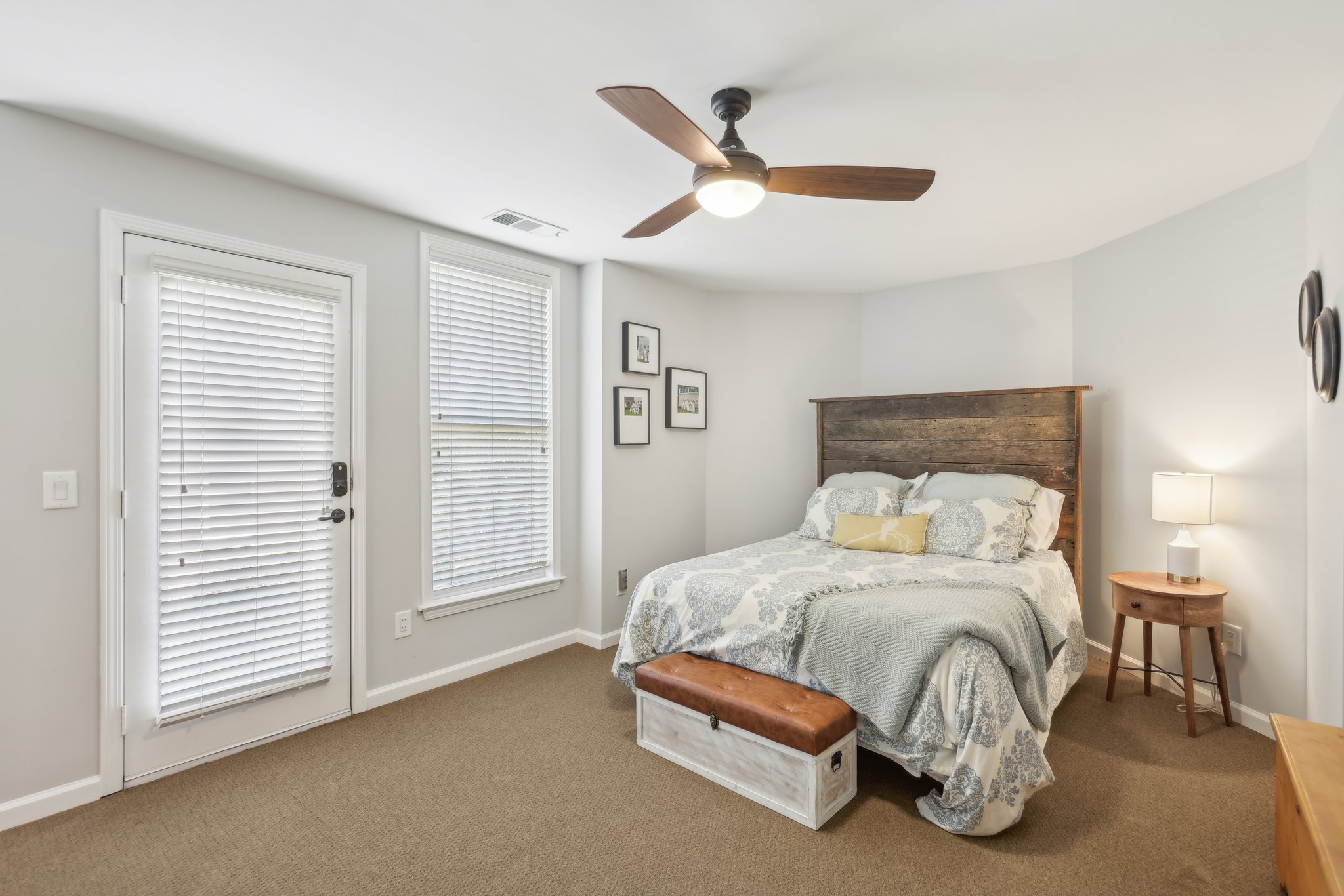 Bedroom with a ceiling fan, wooden headboard bed, patterned bedding, side table with lamp, small art on walls, beige carpet, white door and window with blinds.