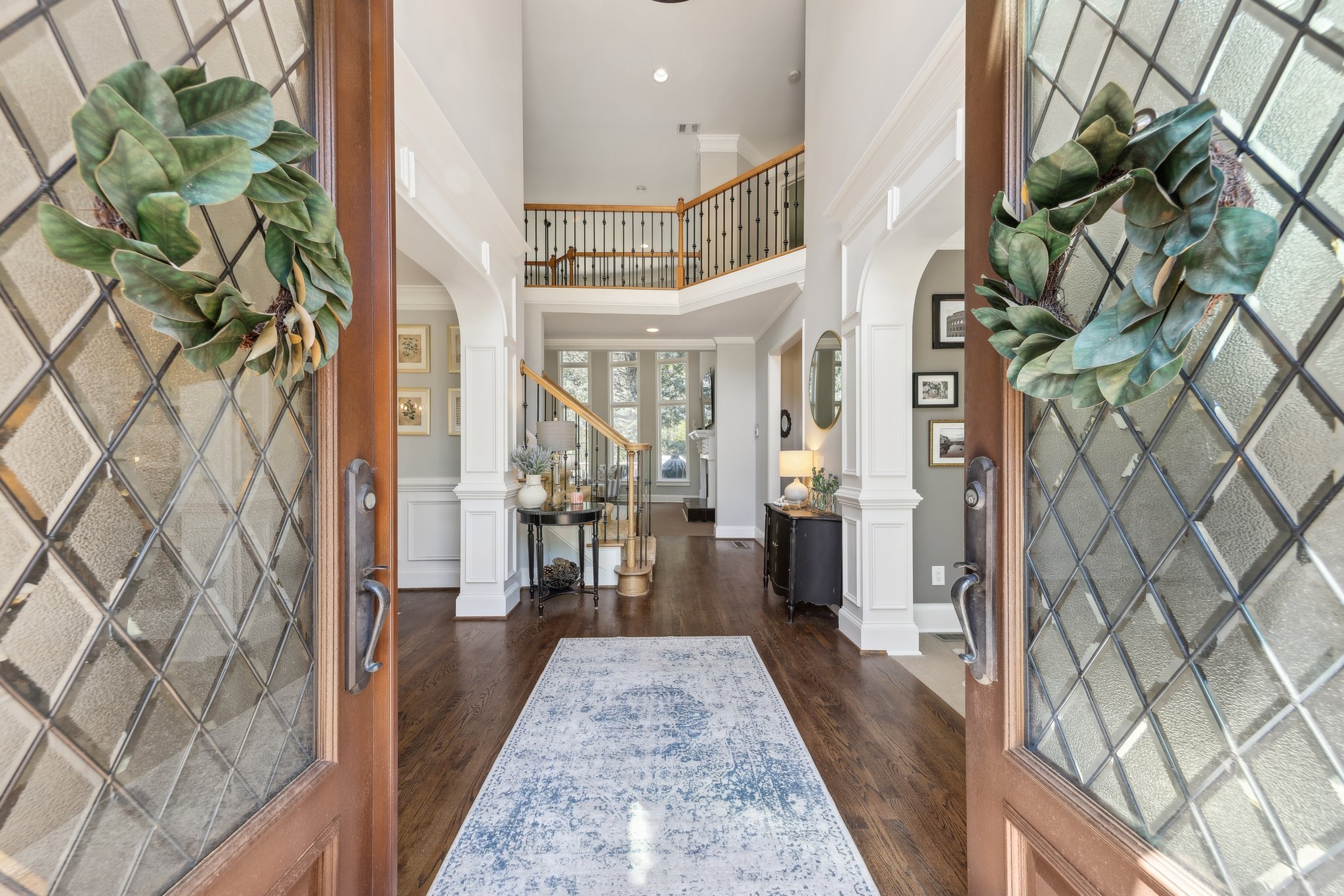 Open entrance to a spacious, elegant home with a wooden front door, adorned with green wreaths. Inside, there's a hallway with a patterned rug, hardwood floors, and decorative molding. A staircase with a wooden railing leads upstairs, and there are t
