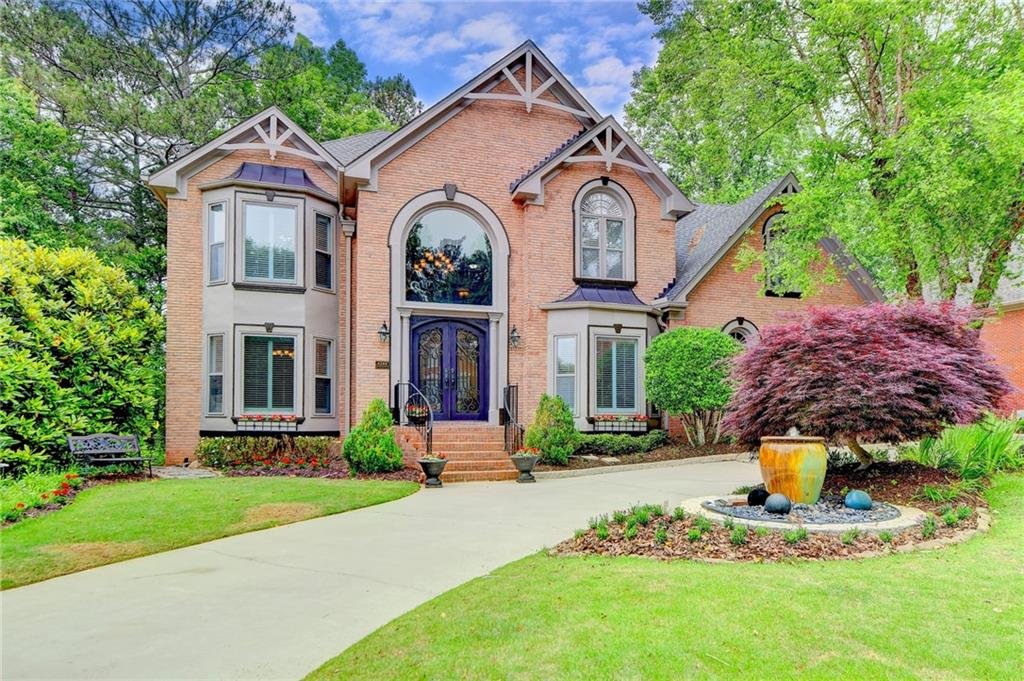 Two-story brick house with large arched window, front porch, and landscaped garden, featuring a decorative urn and plants. Driveway leads to front entrance.